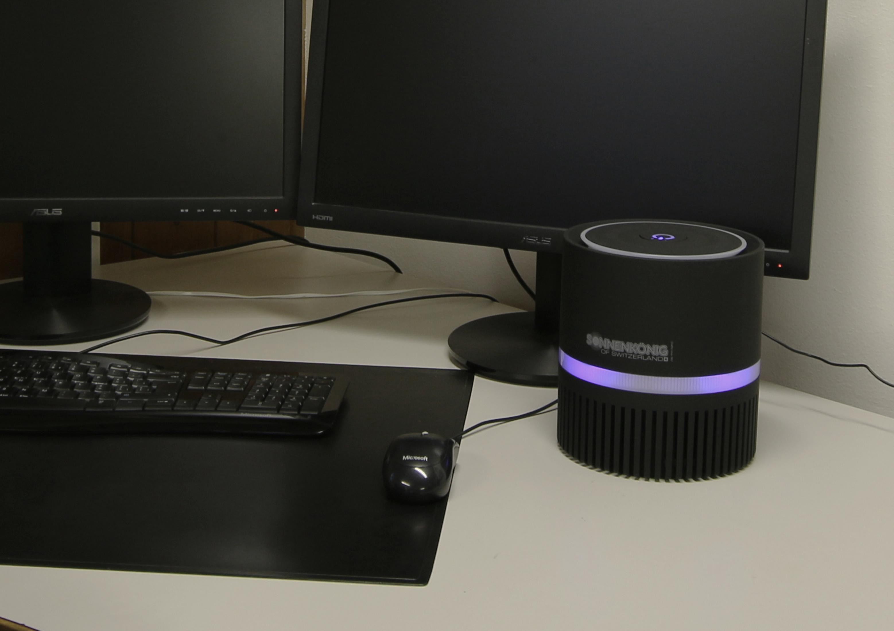 Un purificateur d'air noir sur un bureau blanc avec un clavier, une souris et deux écrans est affiché.