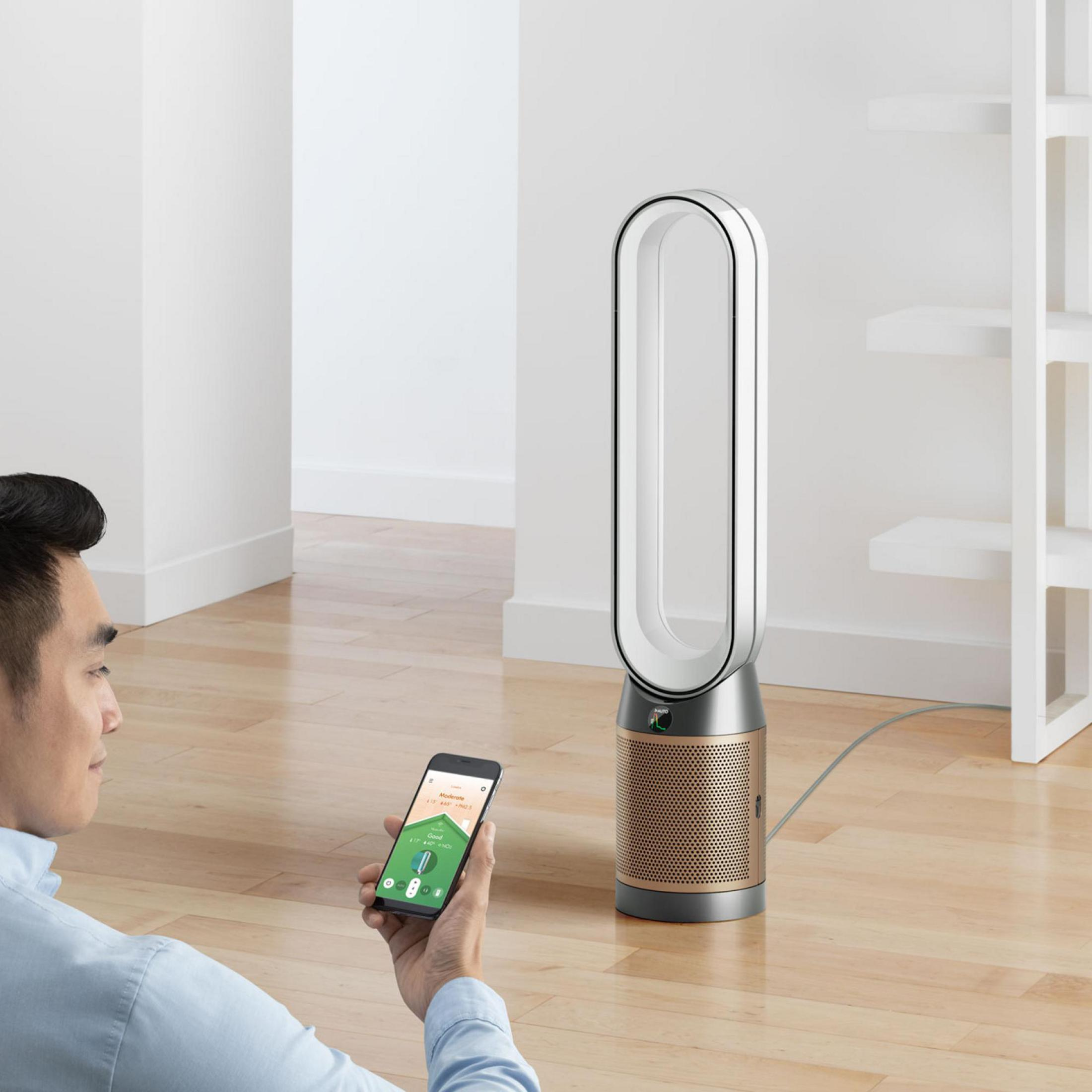 Ein Mann hält ein Telefon neben einem blattlosen Dyson-Ventilator mit einem weißen und kupferfarbenen Farbschema.