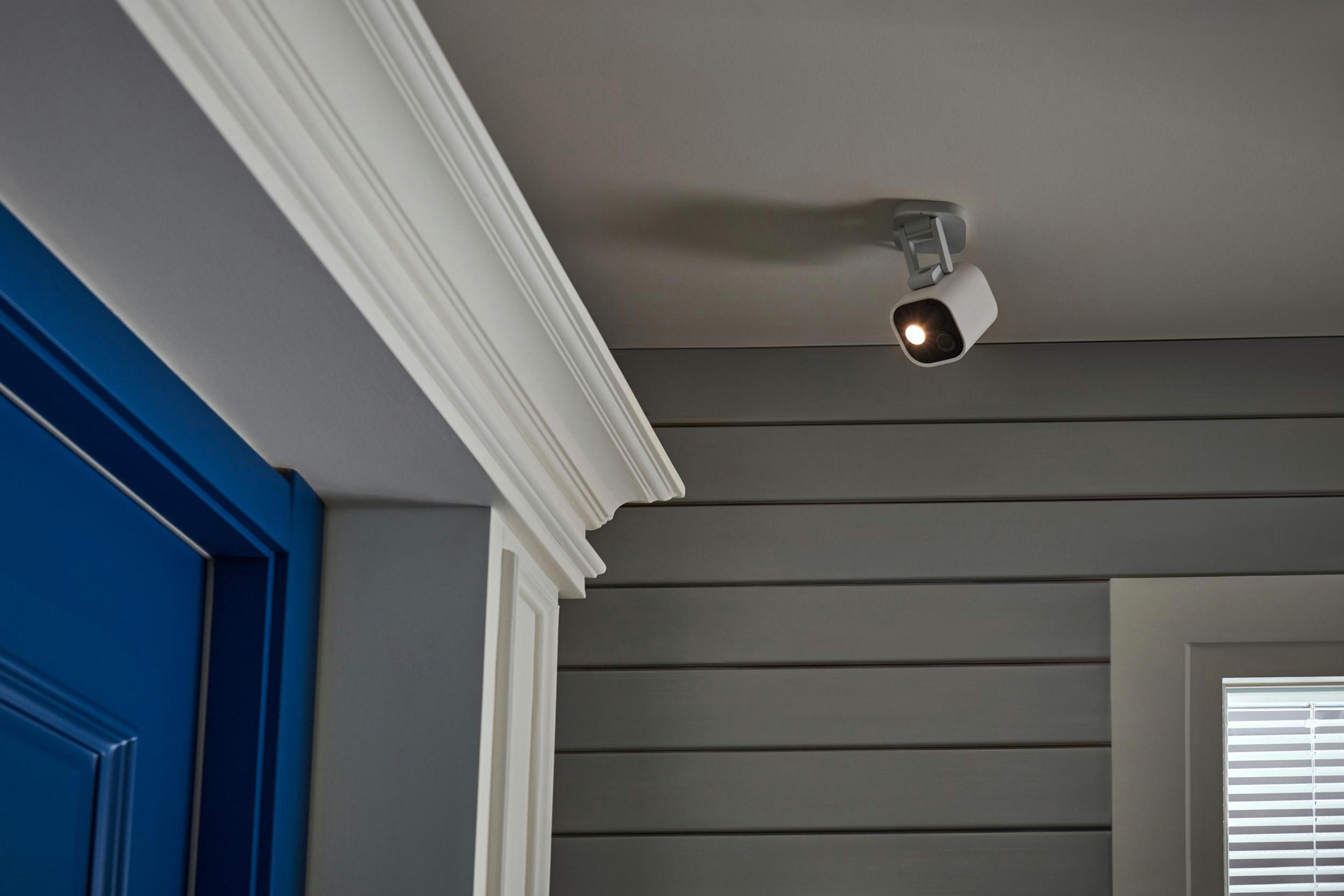 Biała kamera Aqara jest przymocowana do białego sufitu, z niebieskimi drzwiami i szarą ścianą w pobliżu.