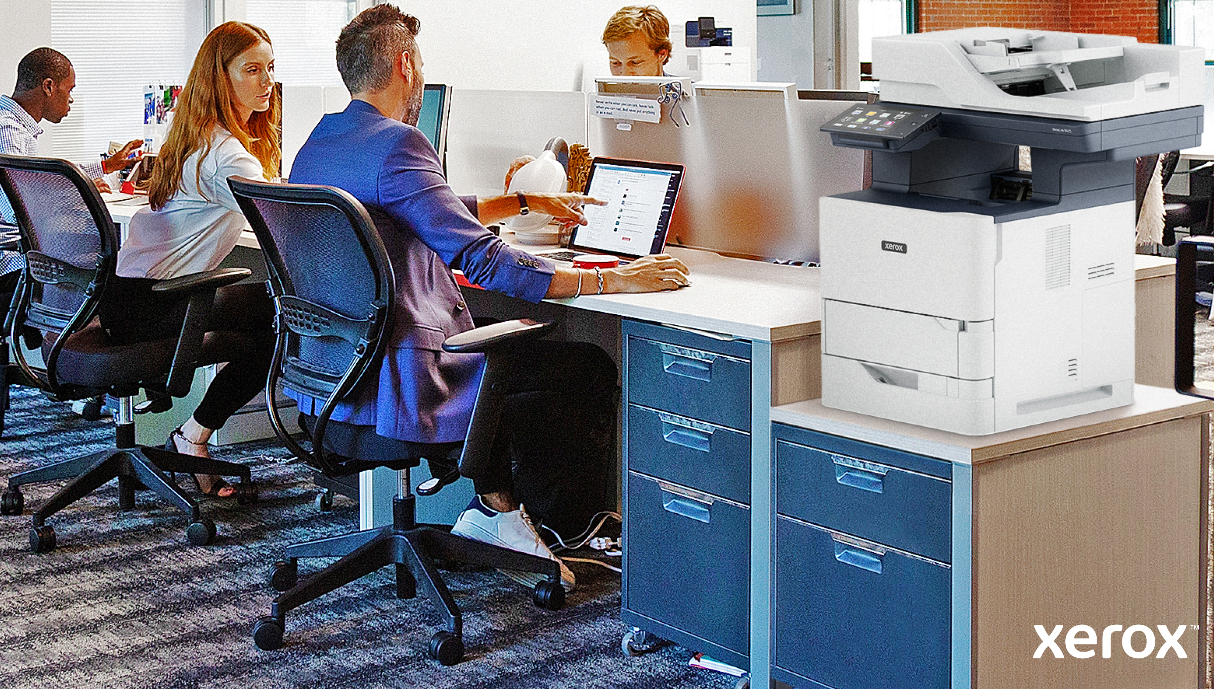 Scena biurowa: ludzie pracują przy biurkach z laptopami. Po prawej stronie stoi duża drukarka Xerox.