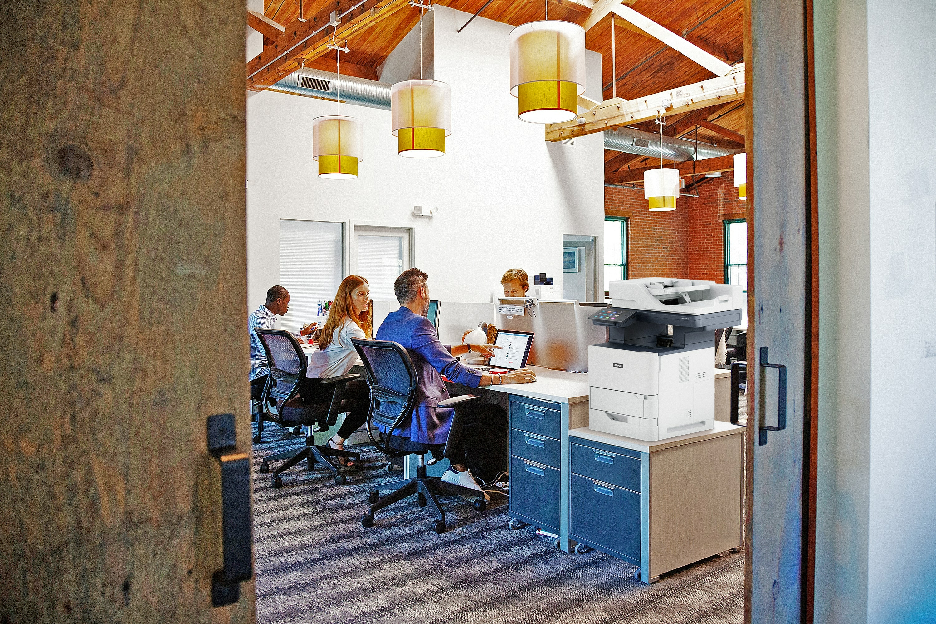 Otoczenie biurowe z pracującymi ludźmi, drukarką Xerox, drewnianymi belkami i wiszącymi lampami.