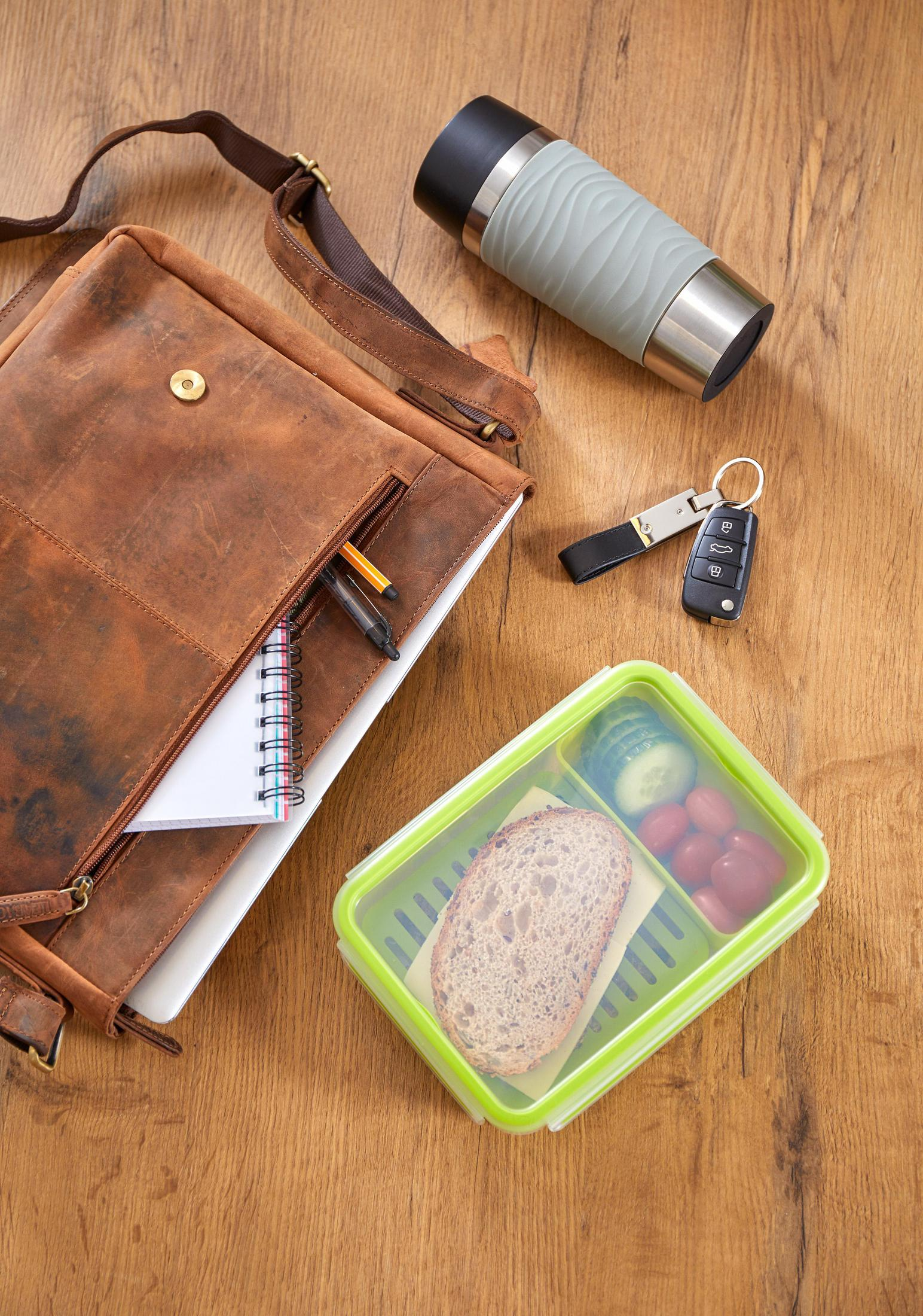 Eine braune Leder-Umhängetasche, Lunchbox, Reisebecher, Schlüssel und Notizbuch auf einem Holztisch.