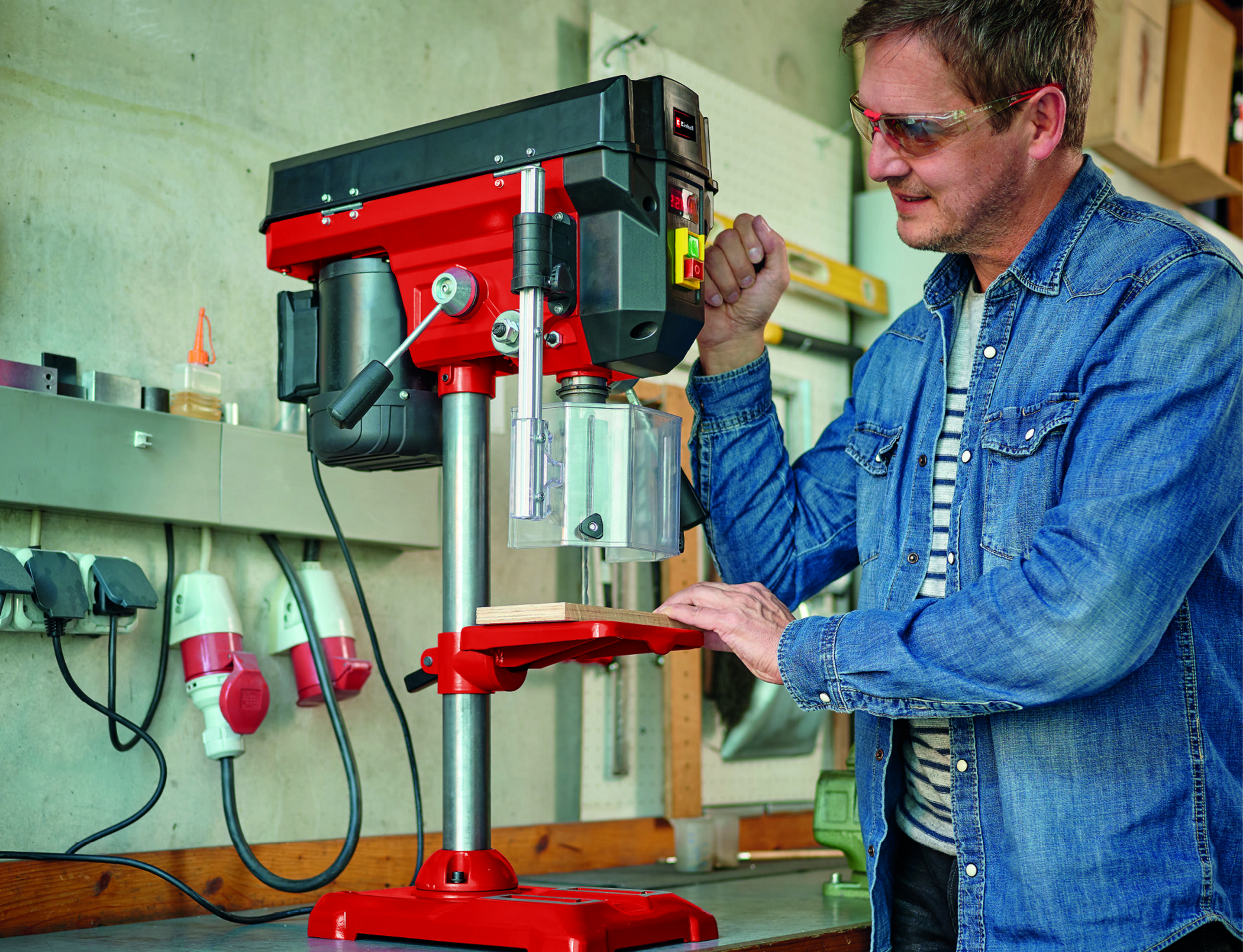 Ein Mann bohrt mit einer roten Tischbohrmaschine ein Loch in ein Stück Holz.
