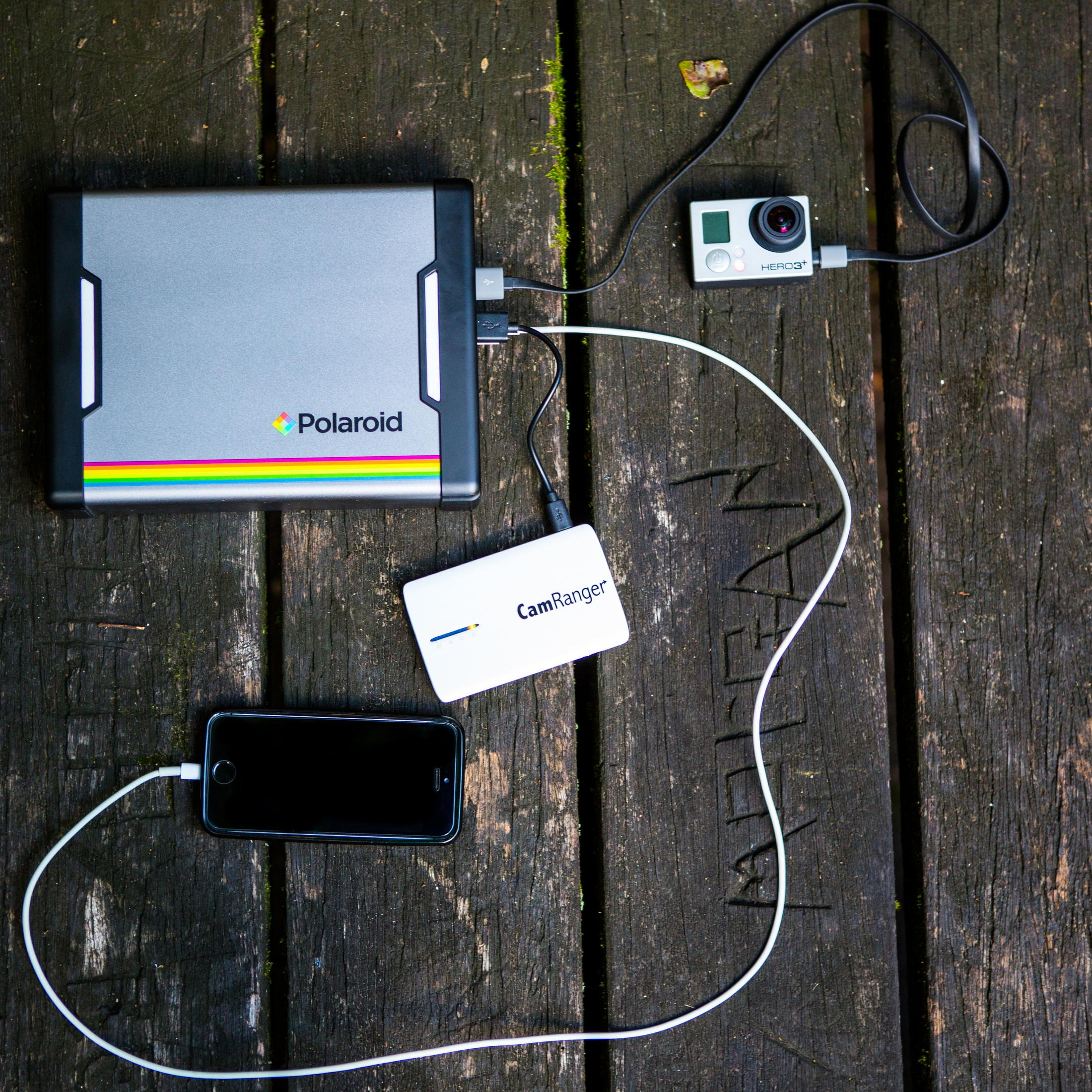 Power bank Polaroid che carica un telefono, una fotocamera e un altro dispositivo sul legno.