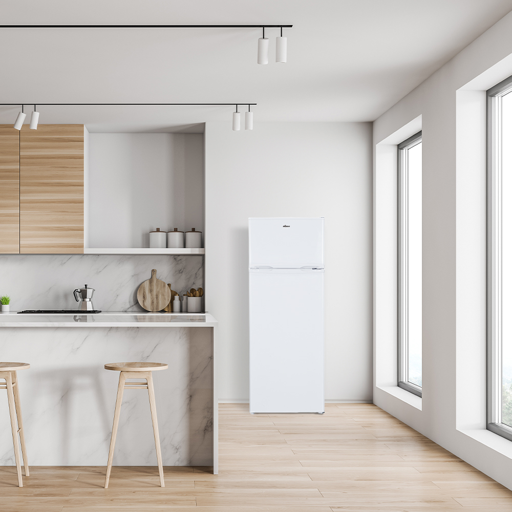 Un refrigerador blanco está colocado en una cocina luminosa con pisos de madera, encimeras de mármol y grandes ventanas.