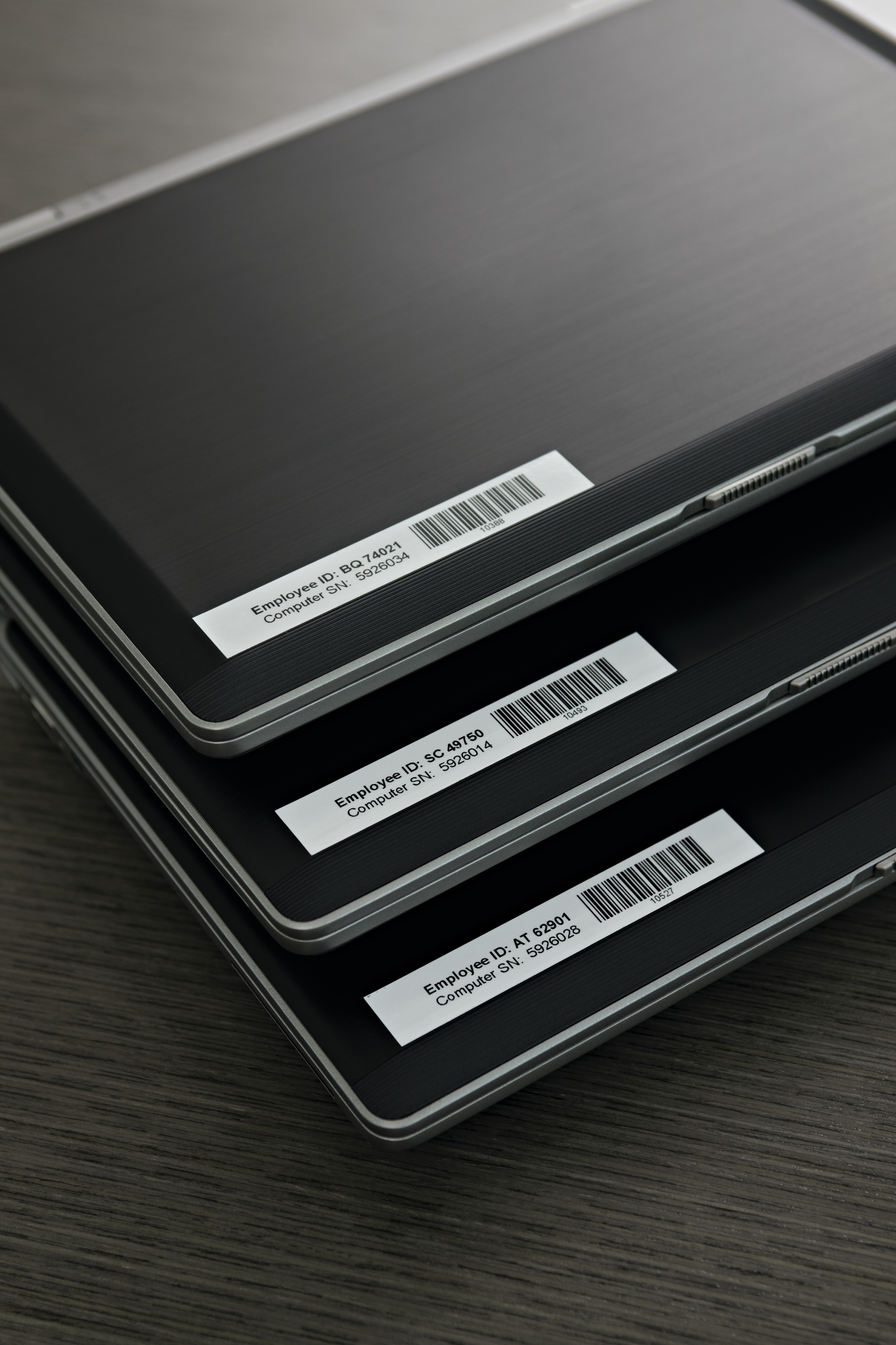 Trzy laptopy ułożone jeden na drugim. Każdy ma naklejkę z identyfikatorem pracownika i numerem seryjnym. Ciemne tło.