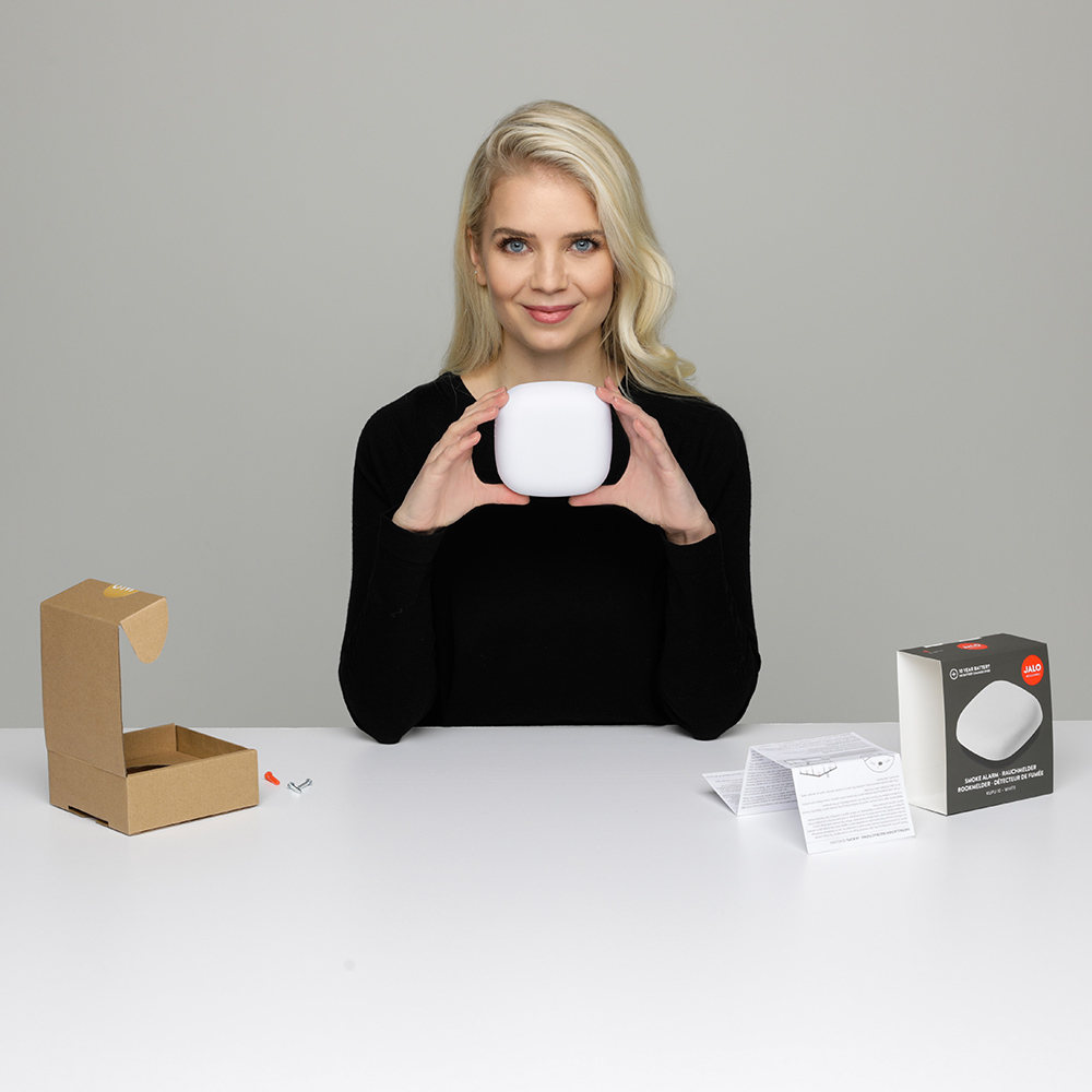 Femme blonde tenant un objet carré blanc et une boîte. Sur une table blanche.