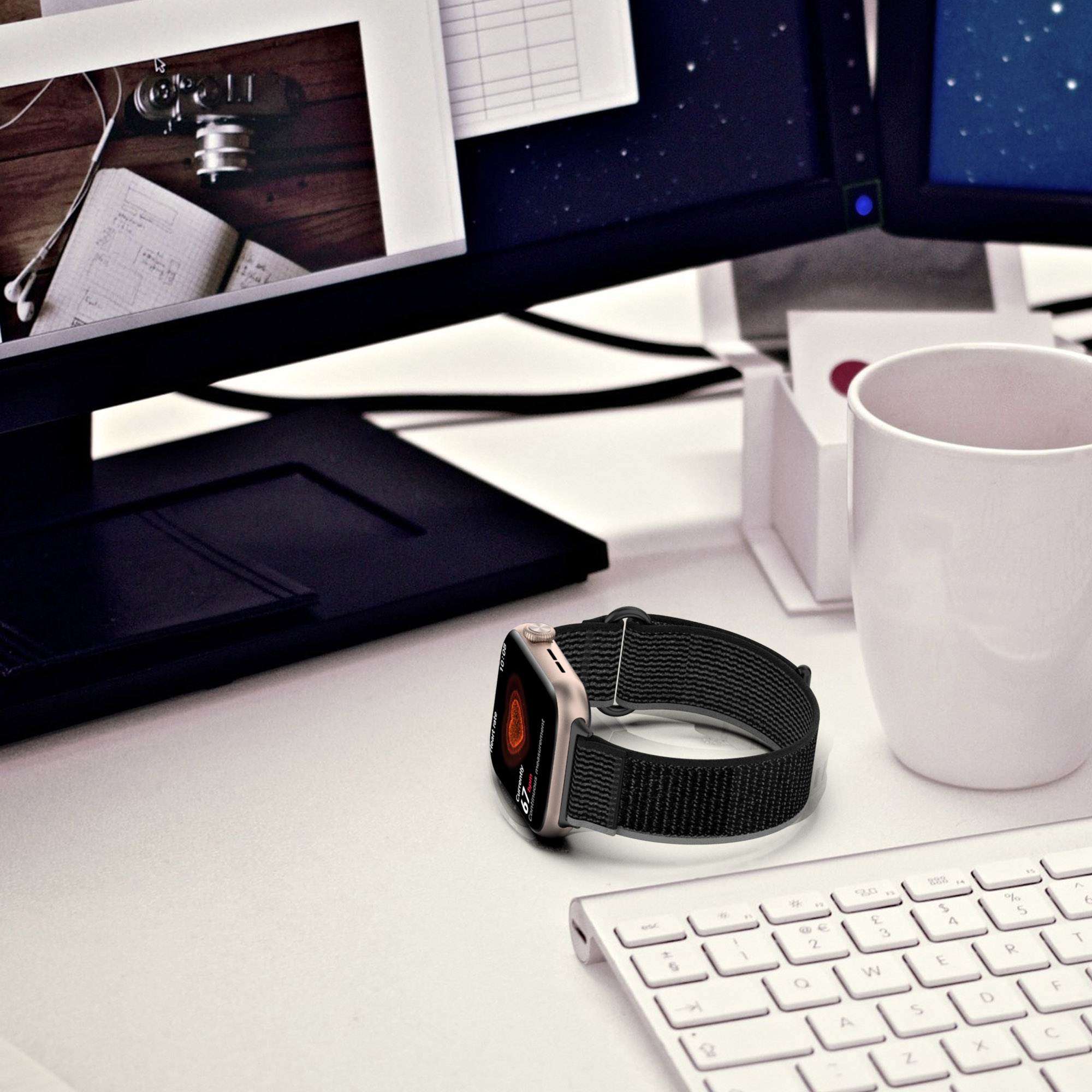 Smartwatch z czarnym paskiem leżący na stole z klawiaturą, monitorem i kubkiem.