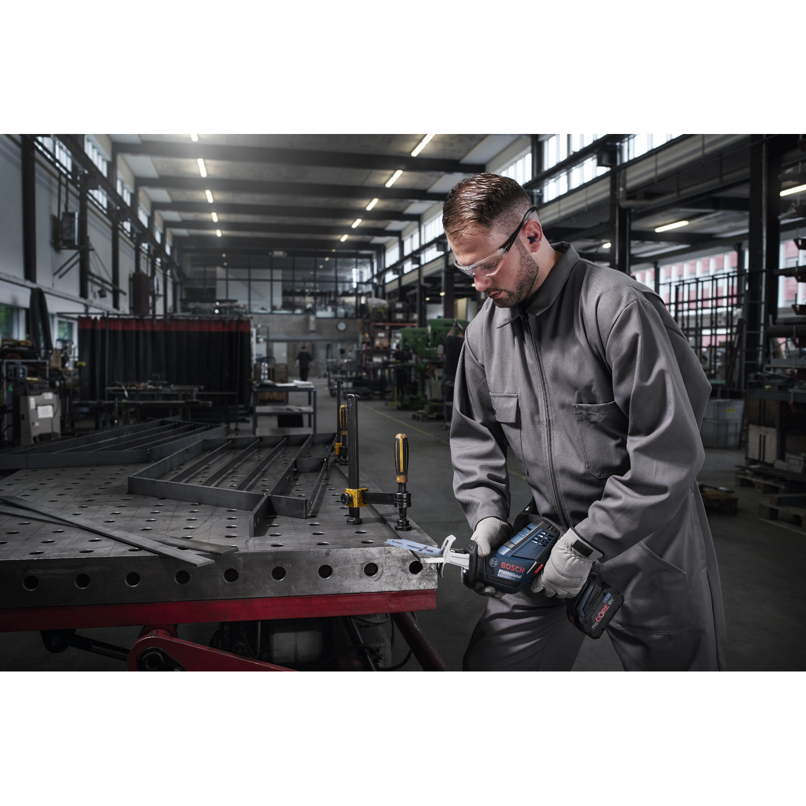 Mężczyzna w szarym kombinezonie używa piły szablastej Bosch w warsztacie. Noszone są okulary ochronne i rękawice.