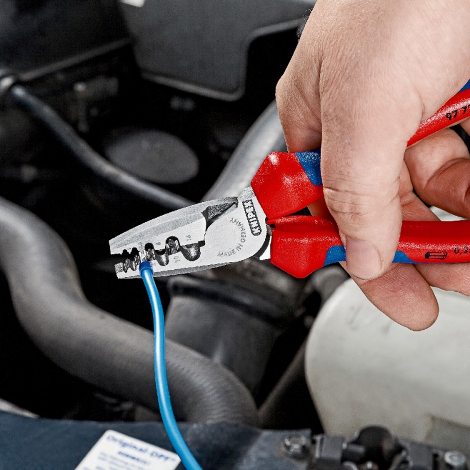 Eine Person verwendet eine Zange, um mit Drähten in einem Motorraum zu arbeiten. Die Zange hat rot-blaue Griffe.