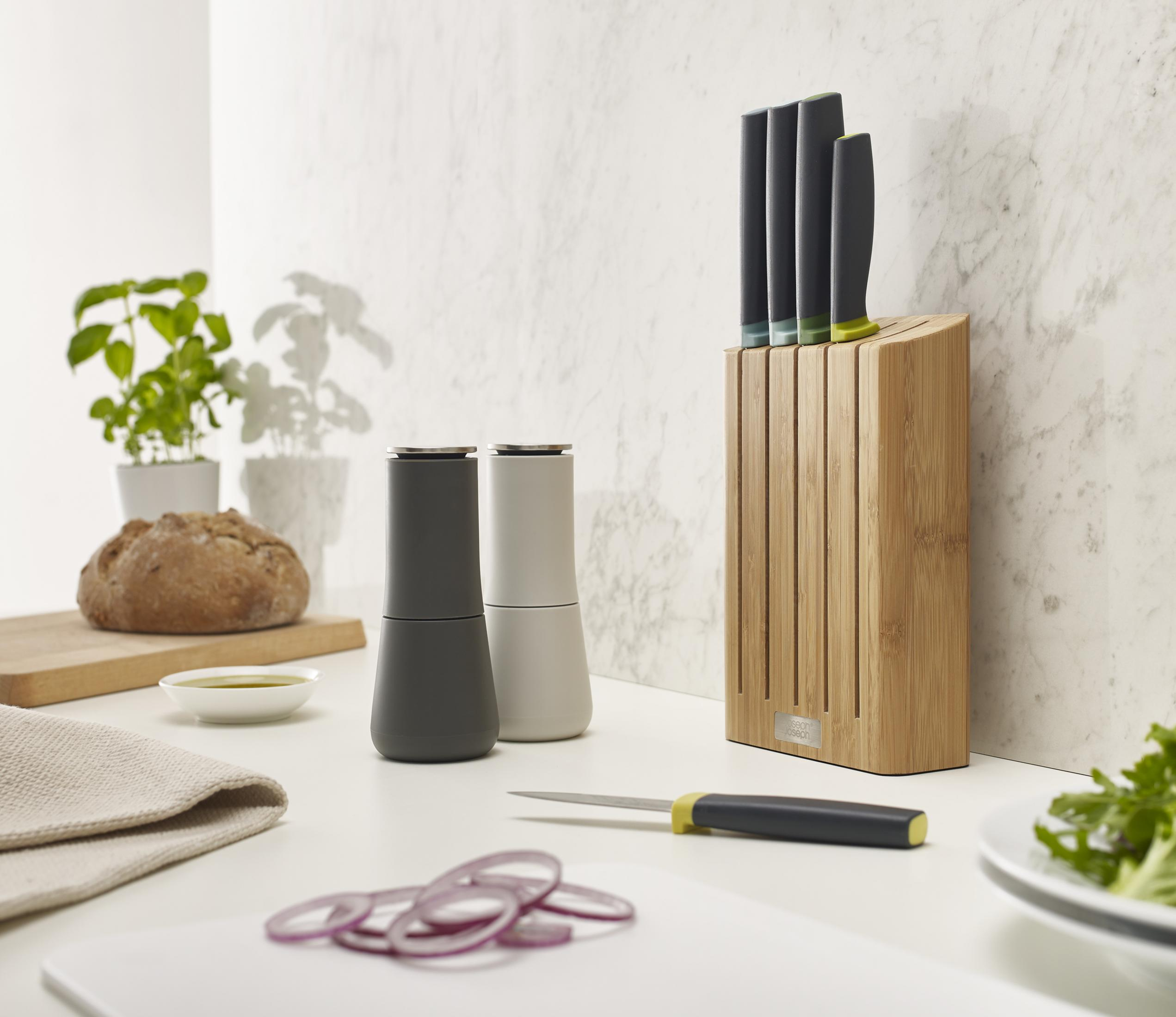 Escena de cocina con cuchillos en un bloque de madera, molinillos, pan y cebollas rebanadas en una encimera blanca.