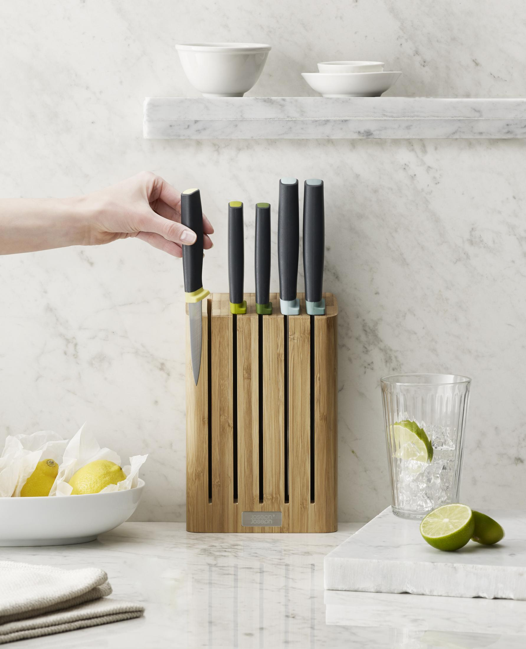 Entorno de cocina con una mano colocando un cuchillo en un bloque de cuchillos de madera, junto a limones, limas y tazones.