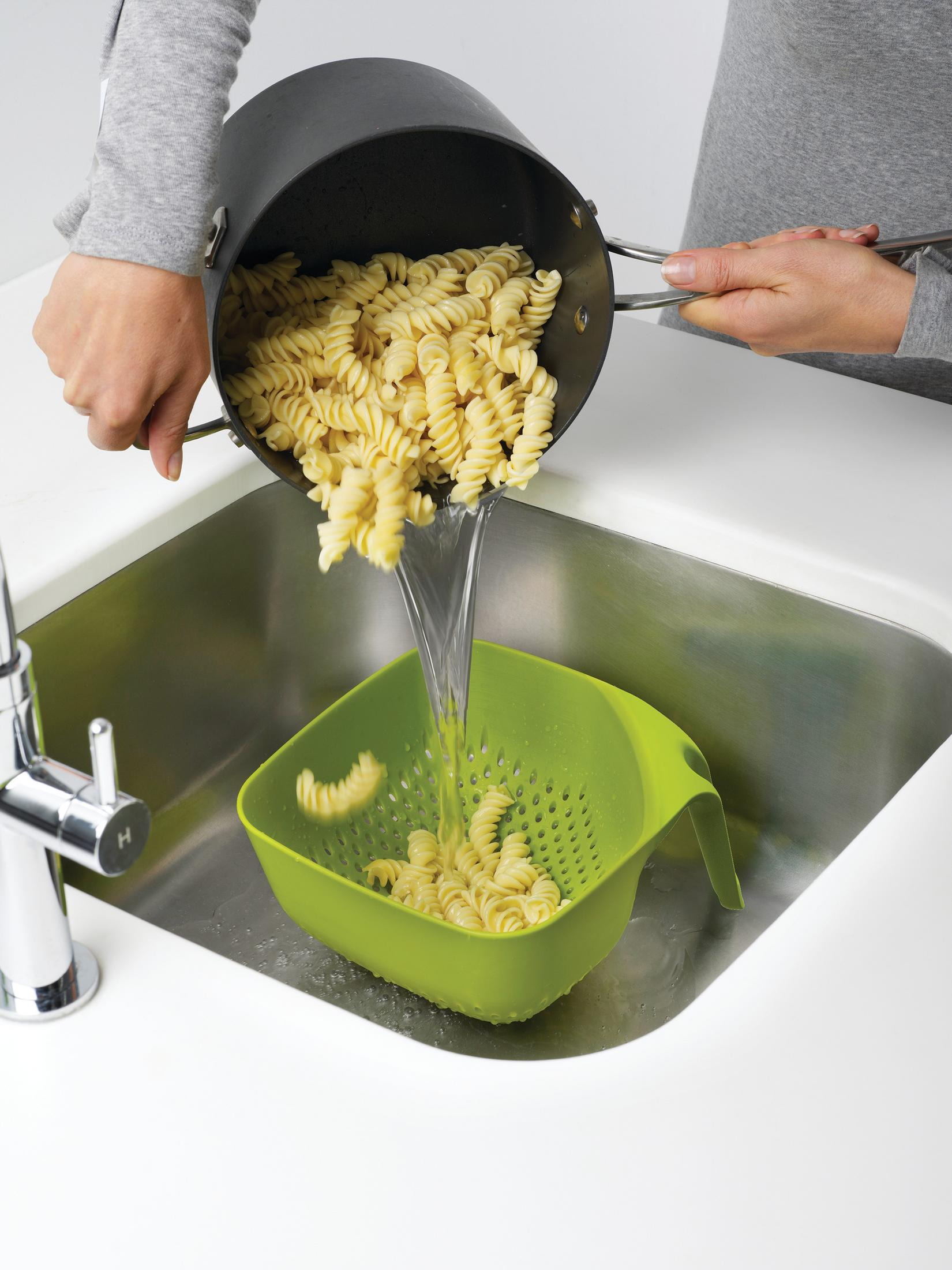 Una persona vierte pasta cocida de una olla negra en un colador verde en un fregadero.