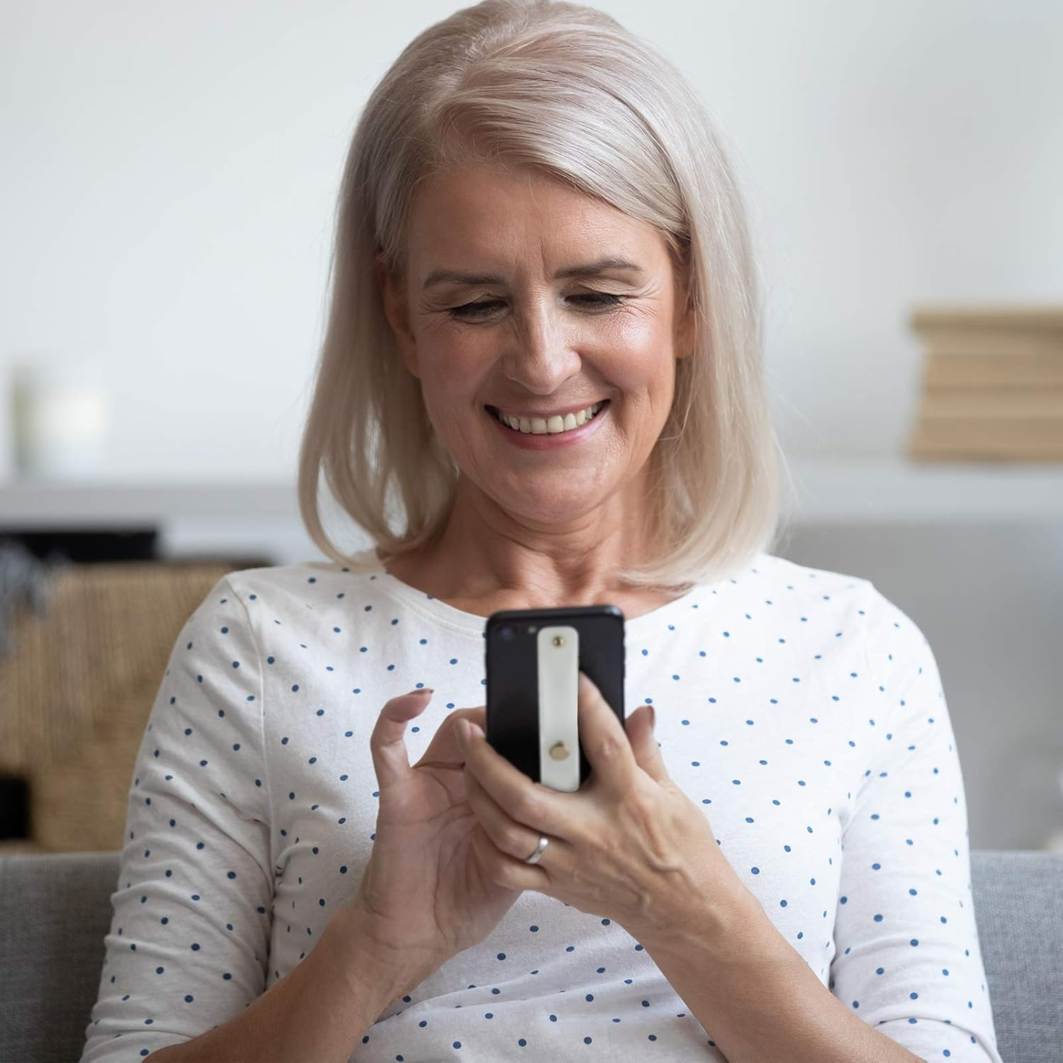 Eine reife Frau lächelt, während sie ein Telefon mit einem weißen Fingergriff hält. Sie trägt ein gepunktetes Hemd.