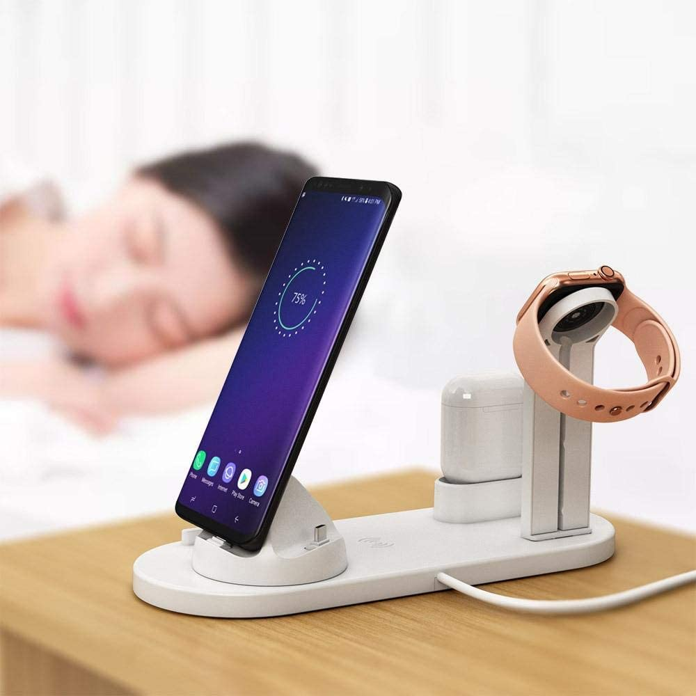 Eine weiße Ladestation mit einem Telefon, einer Smartwatch und Ohrhörern auf einem Holztisch, mit einer verschwommenen Person im Hintergrund.