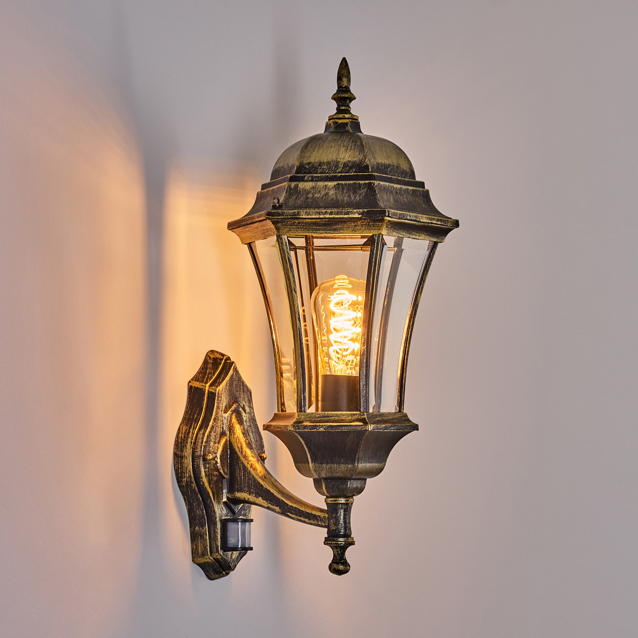 Eine bronzefarbene Außenwandleuchte mit klarem Glas und einem Bewegungssensor an einer weißen Wand, beleuchtet.
