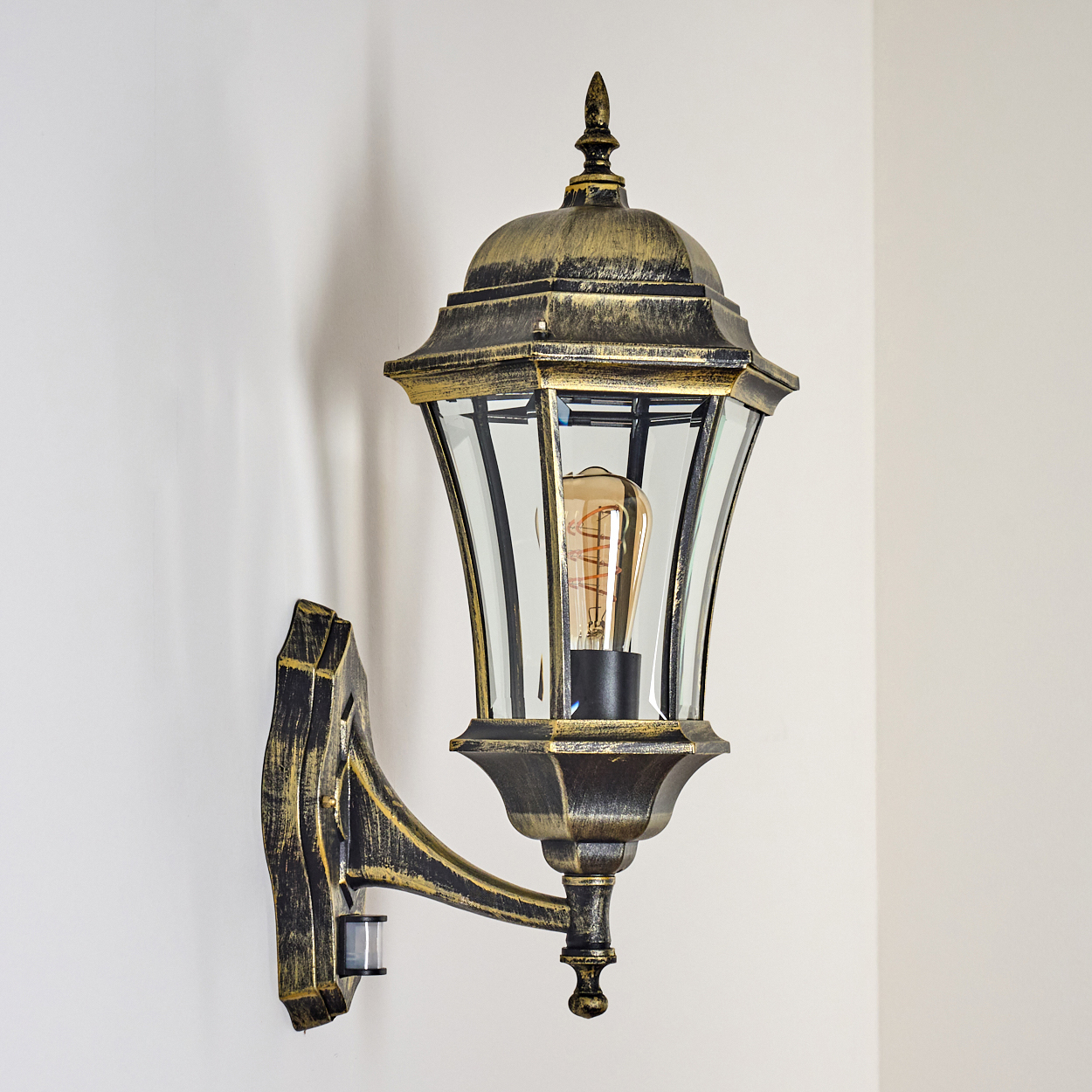 Eine bronzefarbene Außenwandleuchte mit klarem Glas und einem Bewegungssensor an einer weißen Wand.