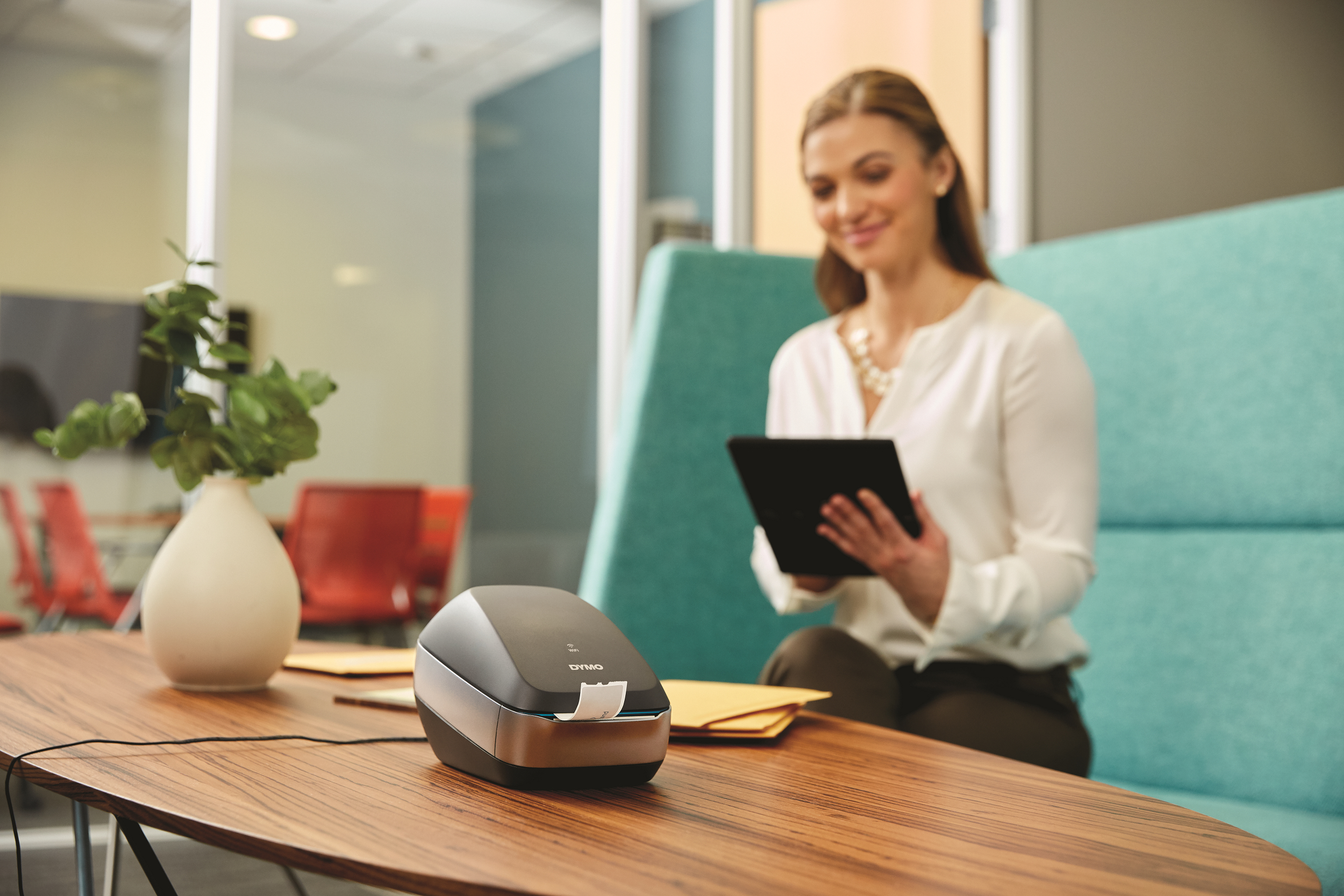 DYMO label printer on a wooden table with a woman holding a tablet in a blurred office setting.