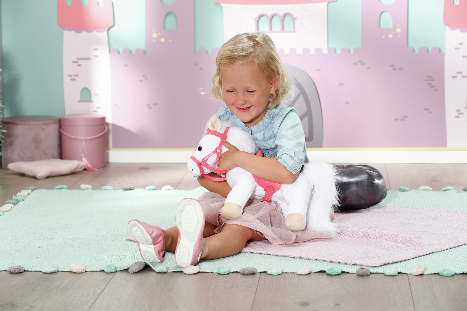 Ein Mädchen sitzt auf dem Boden mit einem Spielzeugpferd. Das Pferd hat einen rosa Sattel und Krone. Sie lächelt.