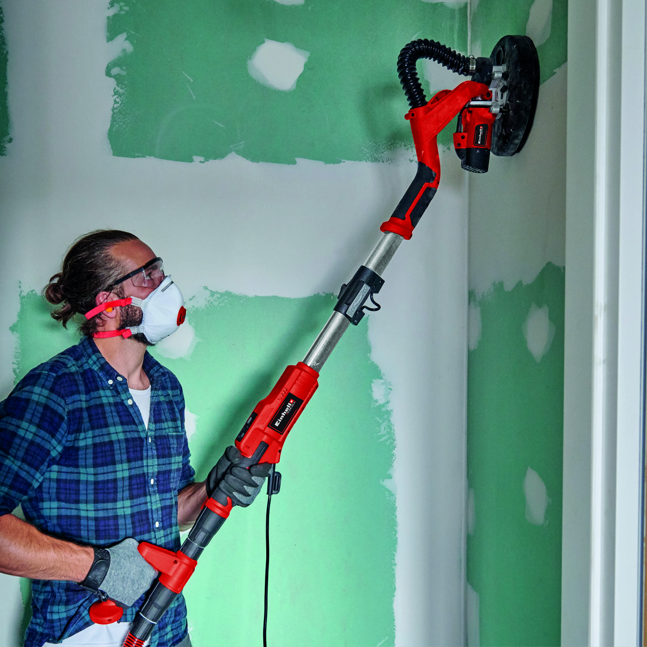 Ein Mann mit Maske benutzt einen roten Trockenbauschleifer an einer Wand.
