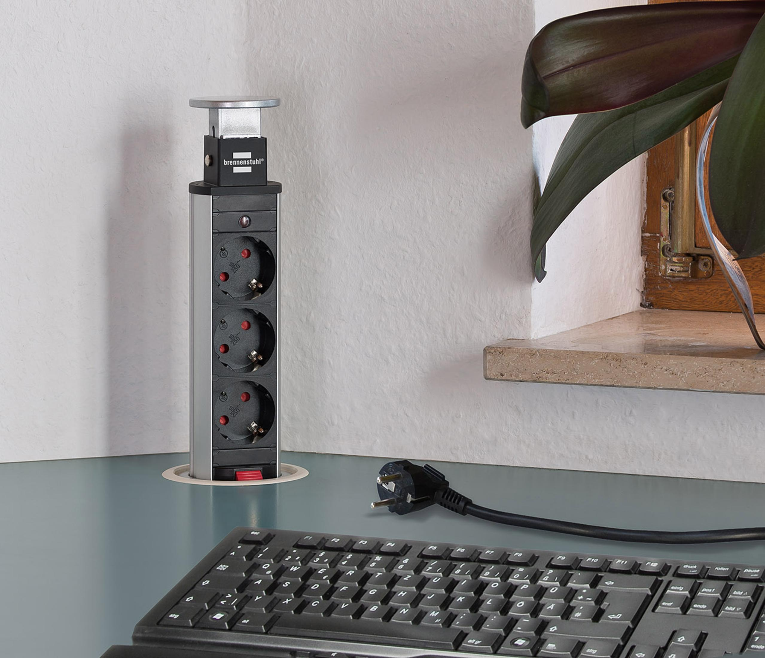 Ein ausfahrbarer Mehrfachstecker mit drei Steckdosen steht auf einem blauen Schreibtisch neben einer Tastatur und einer Pflanze an einem Fenster.