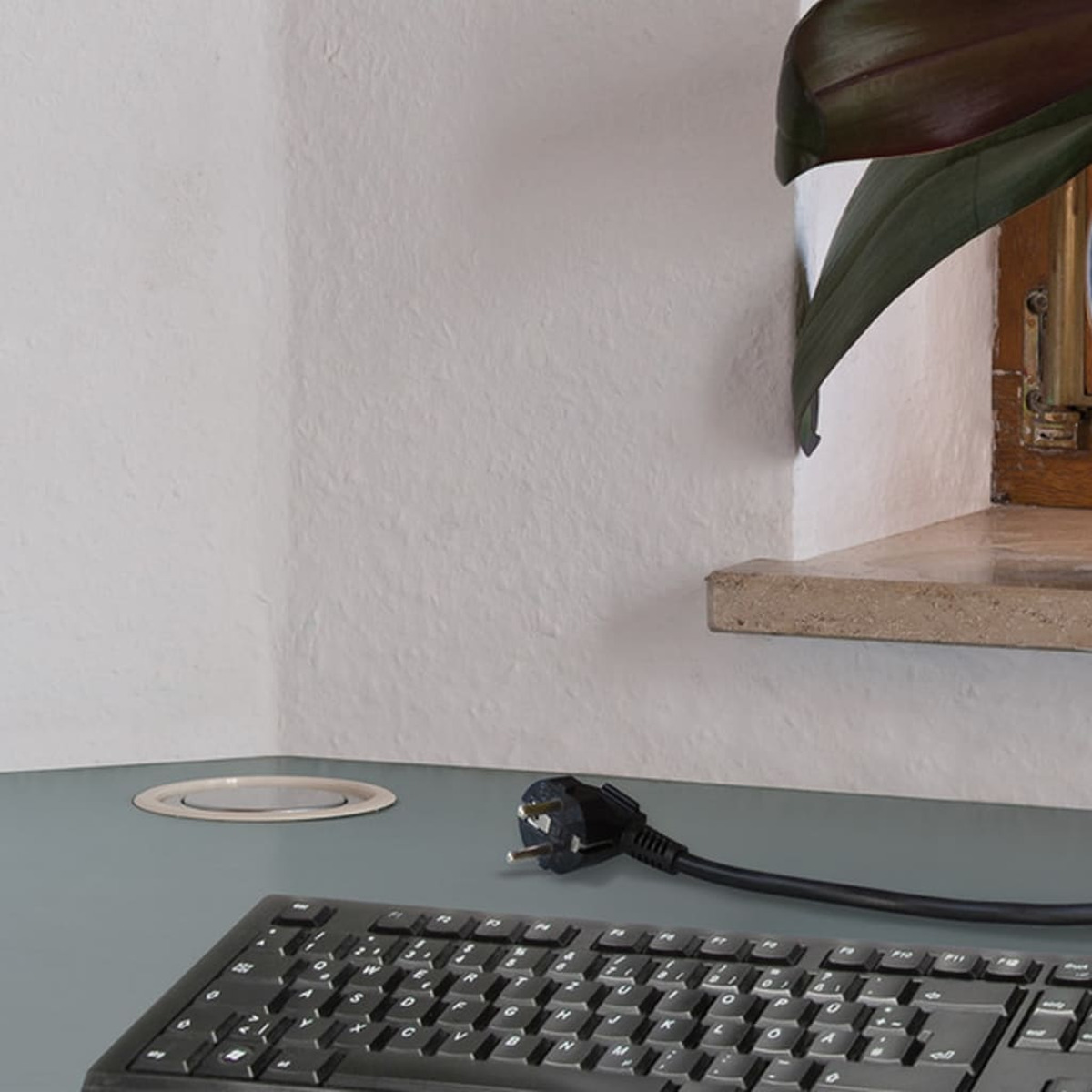 Eine schwarze Tastatur und ein Netzstecker stehen auf einem blauen Schreibtisch, in der Nähe eines Fensters mit einer Pflanze auf der Fensterbank.