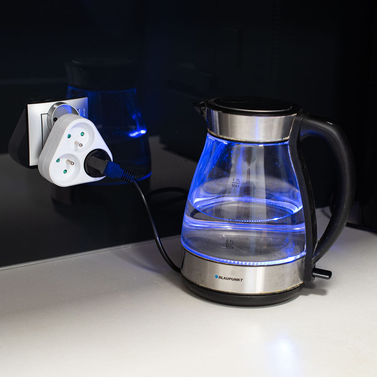 Czajnik podłączony do potrójnego adaptera. Niebieskie światło w środku. Biała powierzchnia.