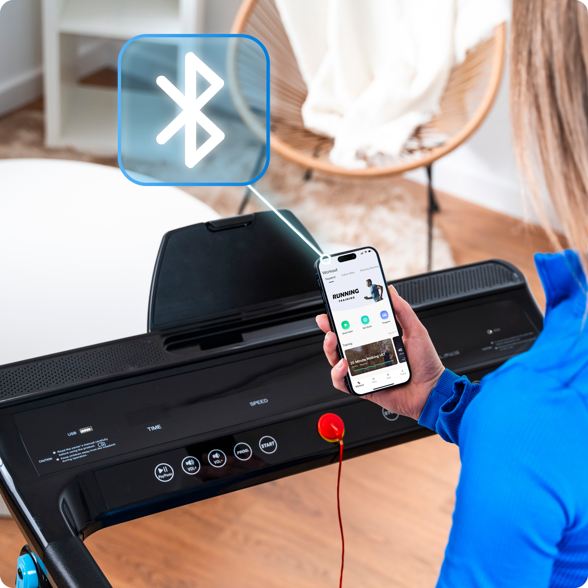 Kobieta trzyma smartfon z aplikacją do biegania, stojąc w pobliżu czarnej bieżni z nałożoną ikoną bluetooth.