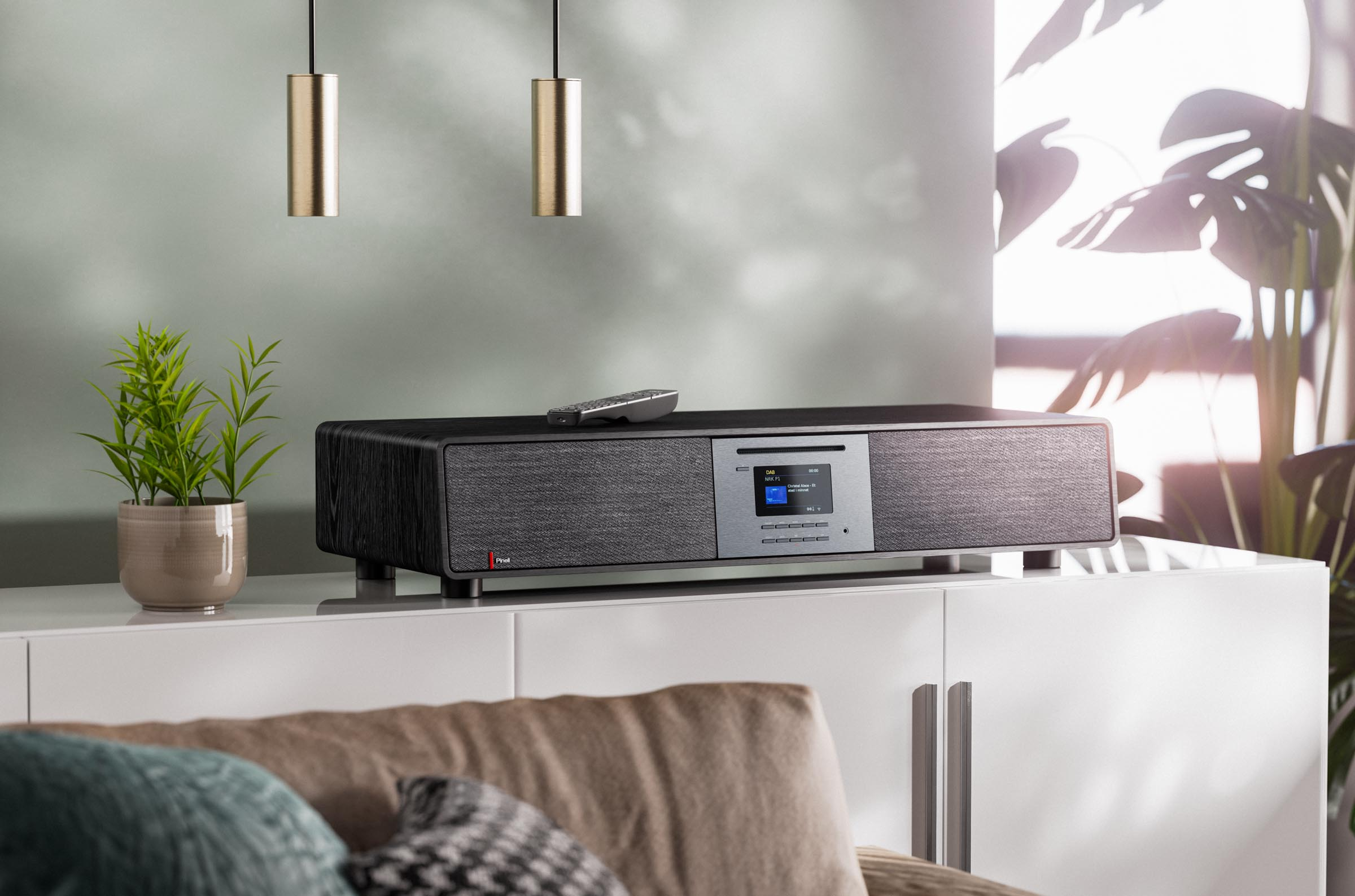 A black audio device on a white cabinet. The scene includes plants, pendant lights and a remote.