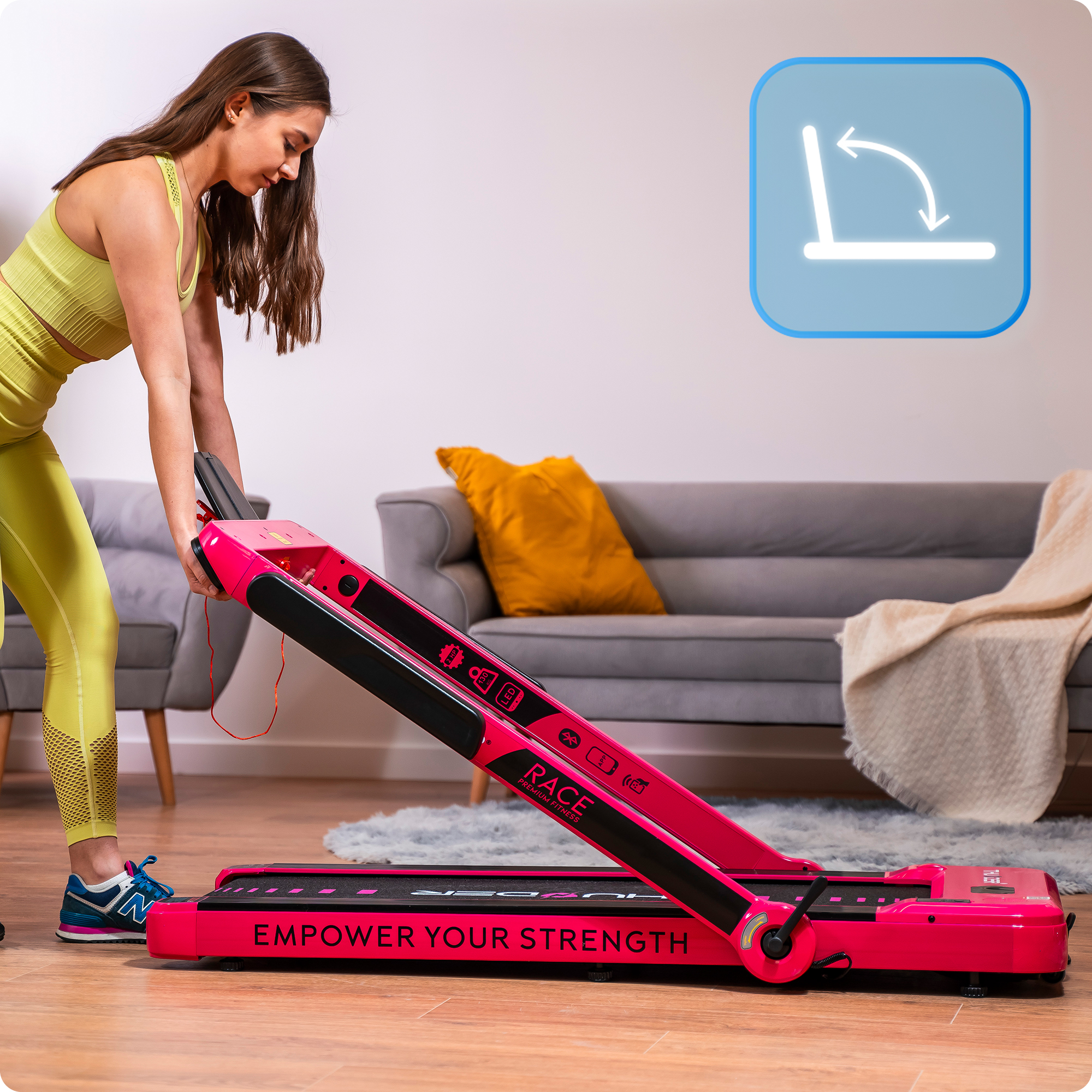 Kobieta składa różową bieżnię w salonie. Bieżnia ma czarne akcenty i napis „EMPOWER YOUR STRENGTH”.