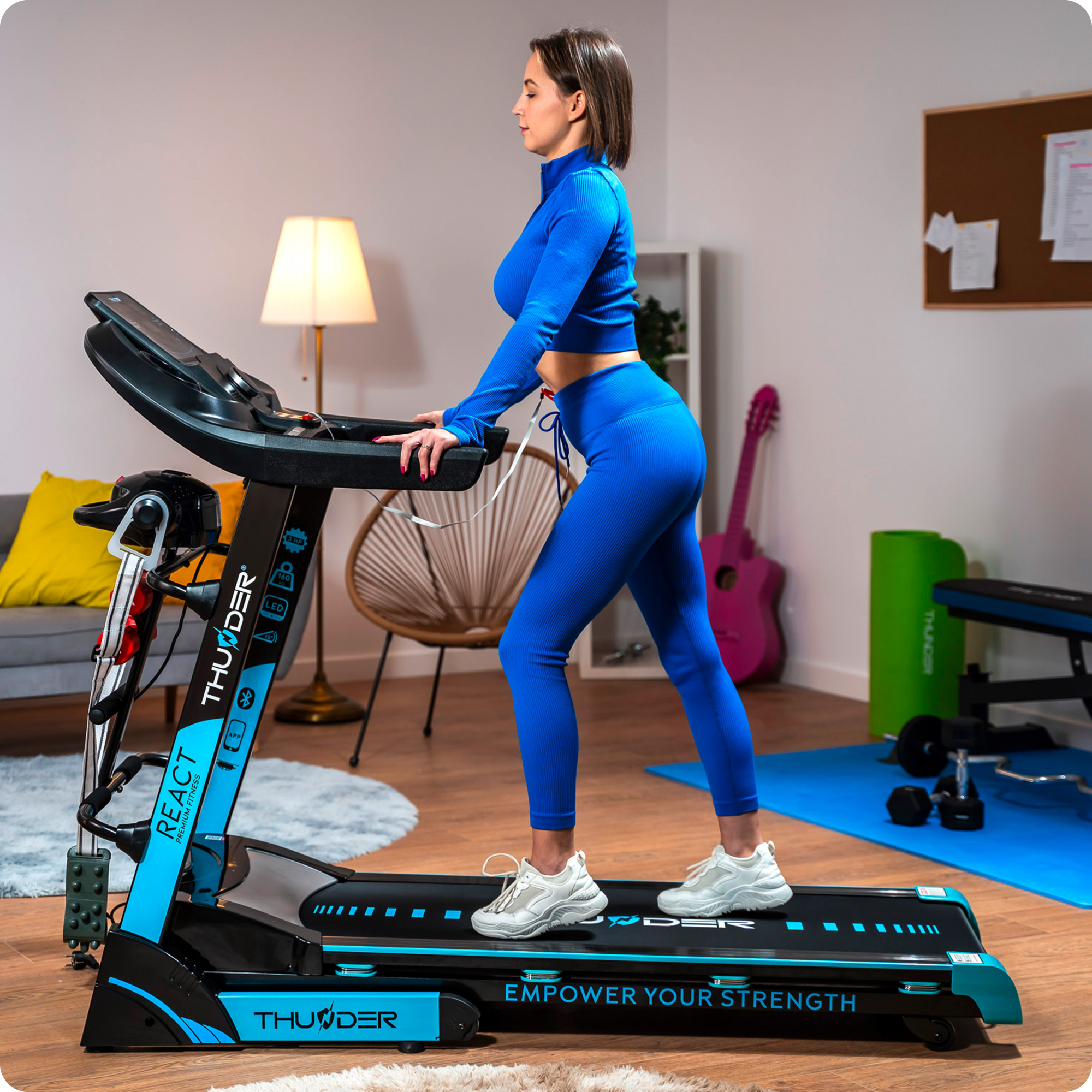 Kobieta w niebieskim stroju do ćwiczeń korzysta z czarno-niebieskiej bieżni w dobrze oświetlonym pomieszczeniu. Fitness i ćwiczenia w domu.