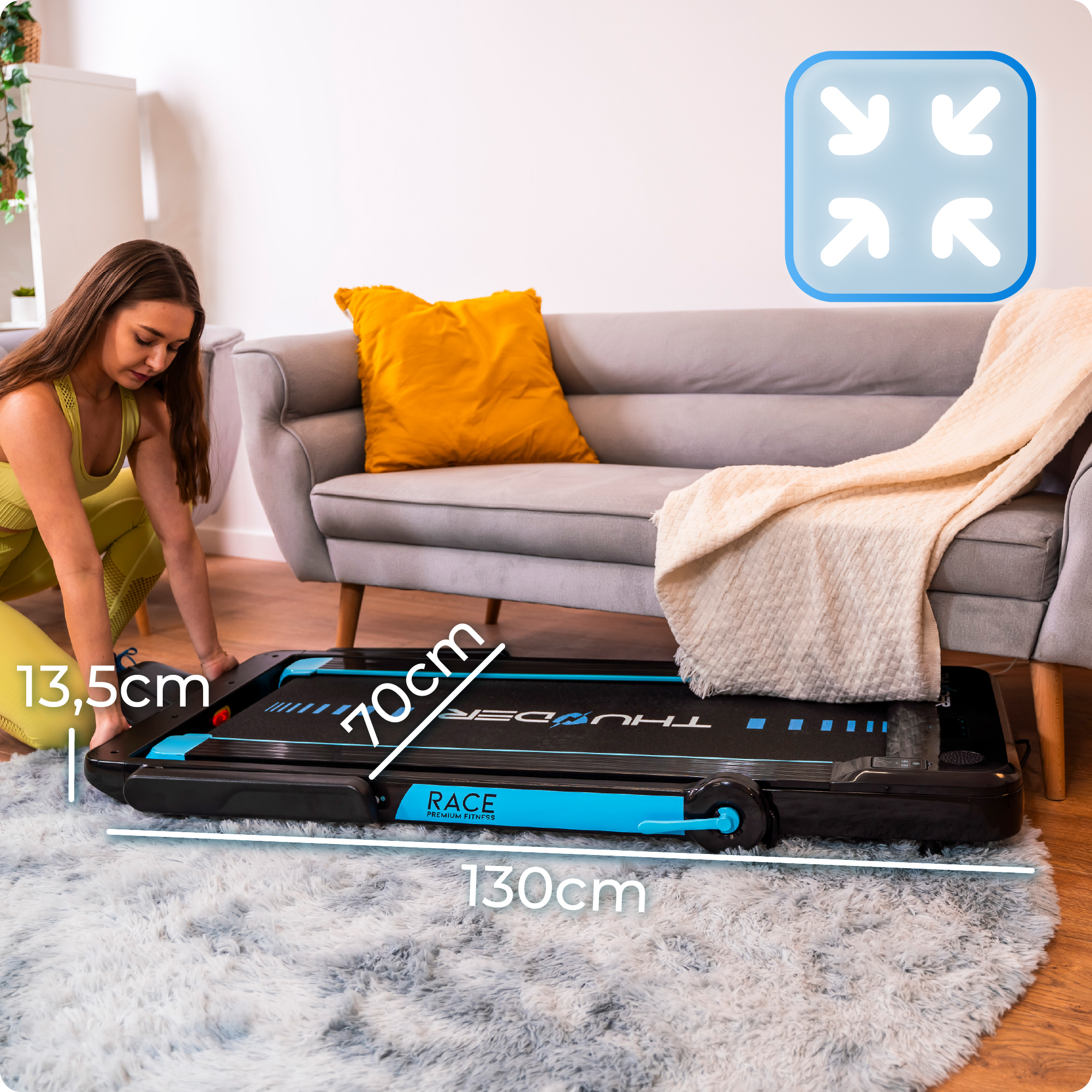 Kobieta reguluje złożoną czarno-niebieską bieżnię na włochatym dywanie w salonie z szarą sofą.