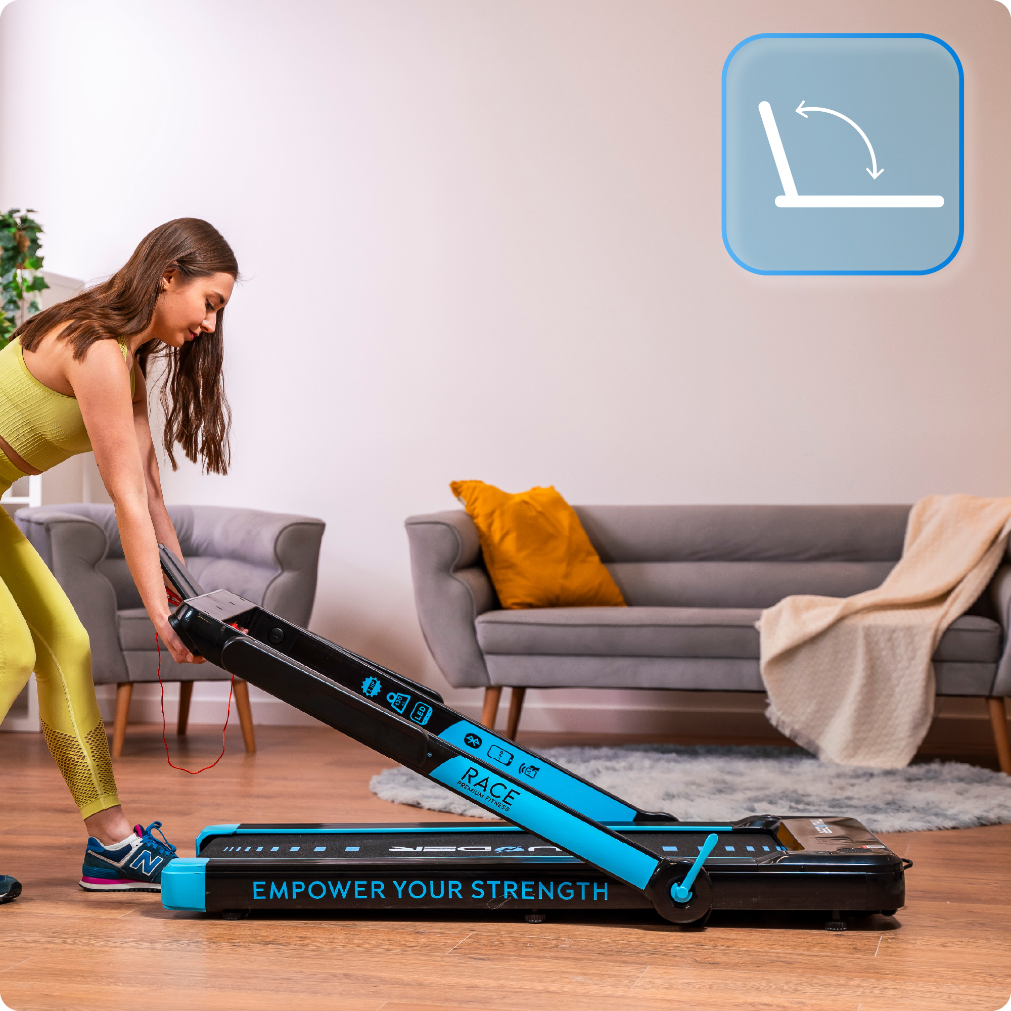 Kobieta składa czarno-niebieską bieżnię. Szara sofa i żółta poduszka w tle.