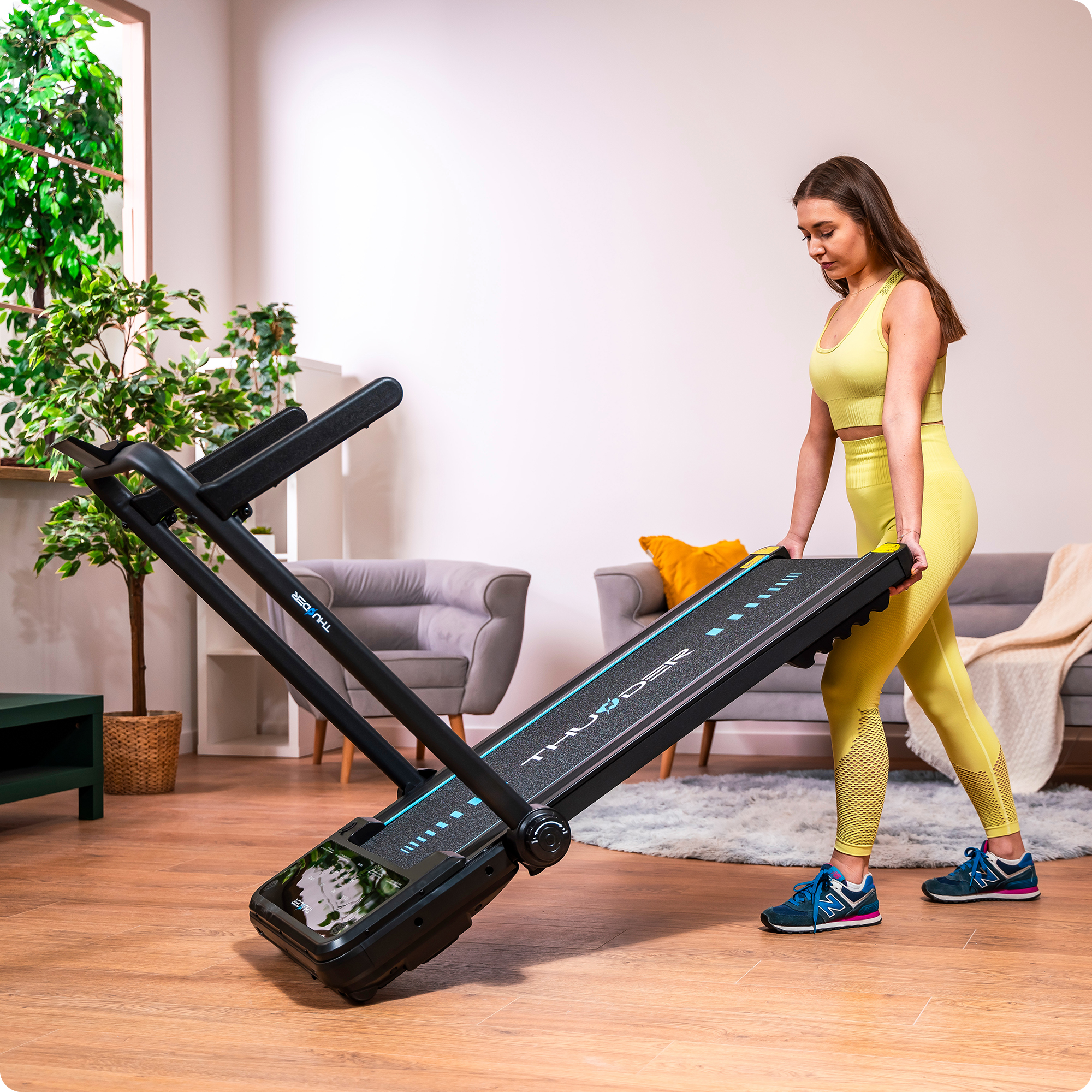 Kobieta podnosi czarną bieżnię. Żółty strój. Szara sofa i roślina w tle.