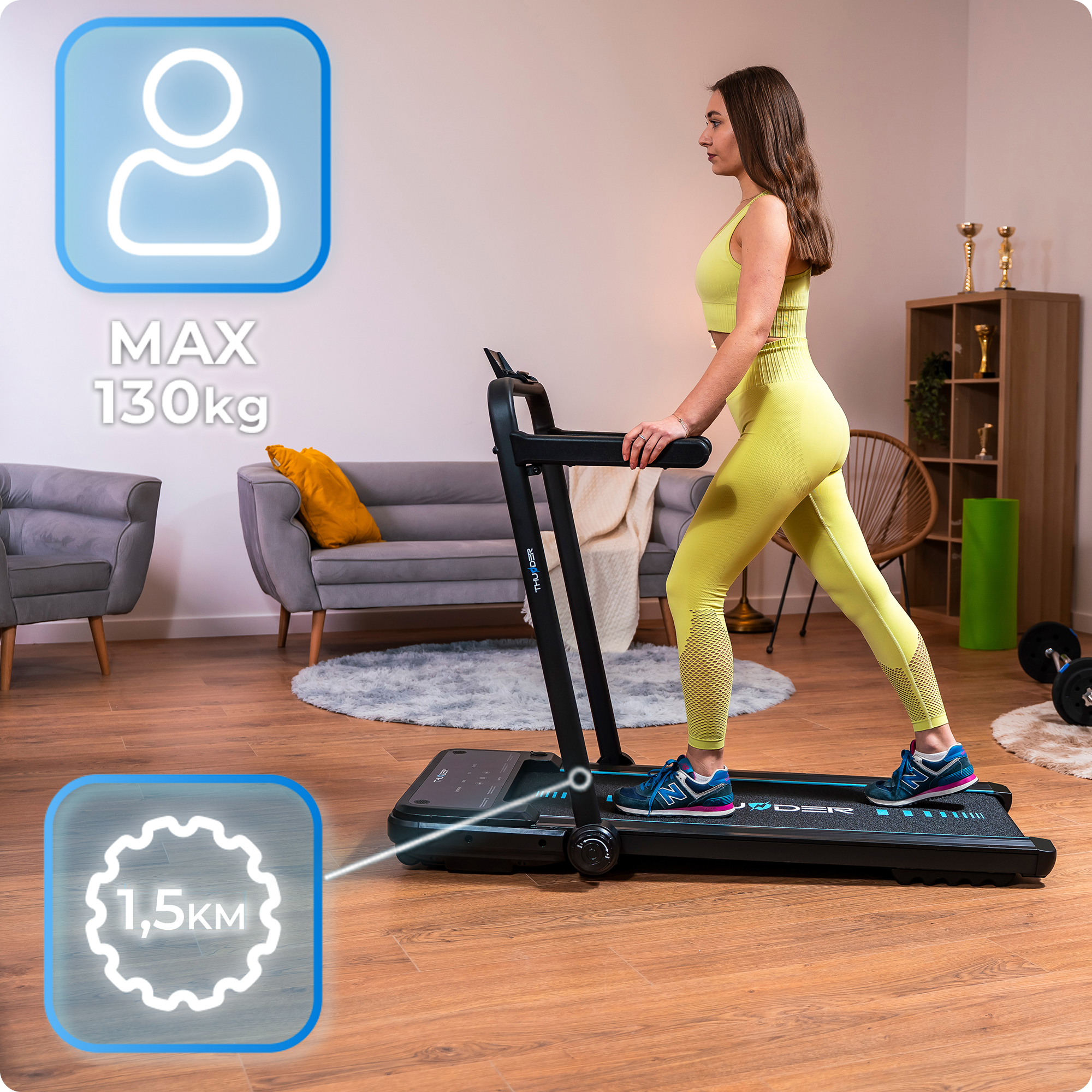 Kobieta idąca po czarnej bieżni. Żółty strój. Szara sofa i trofea w tle.