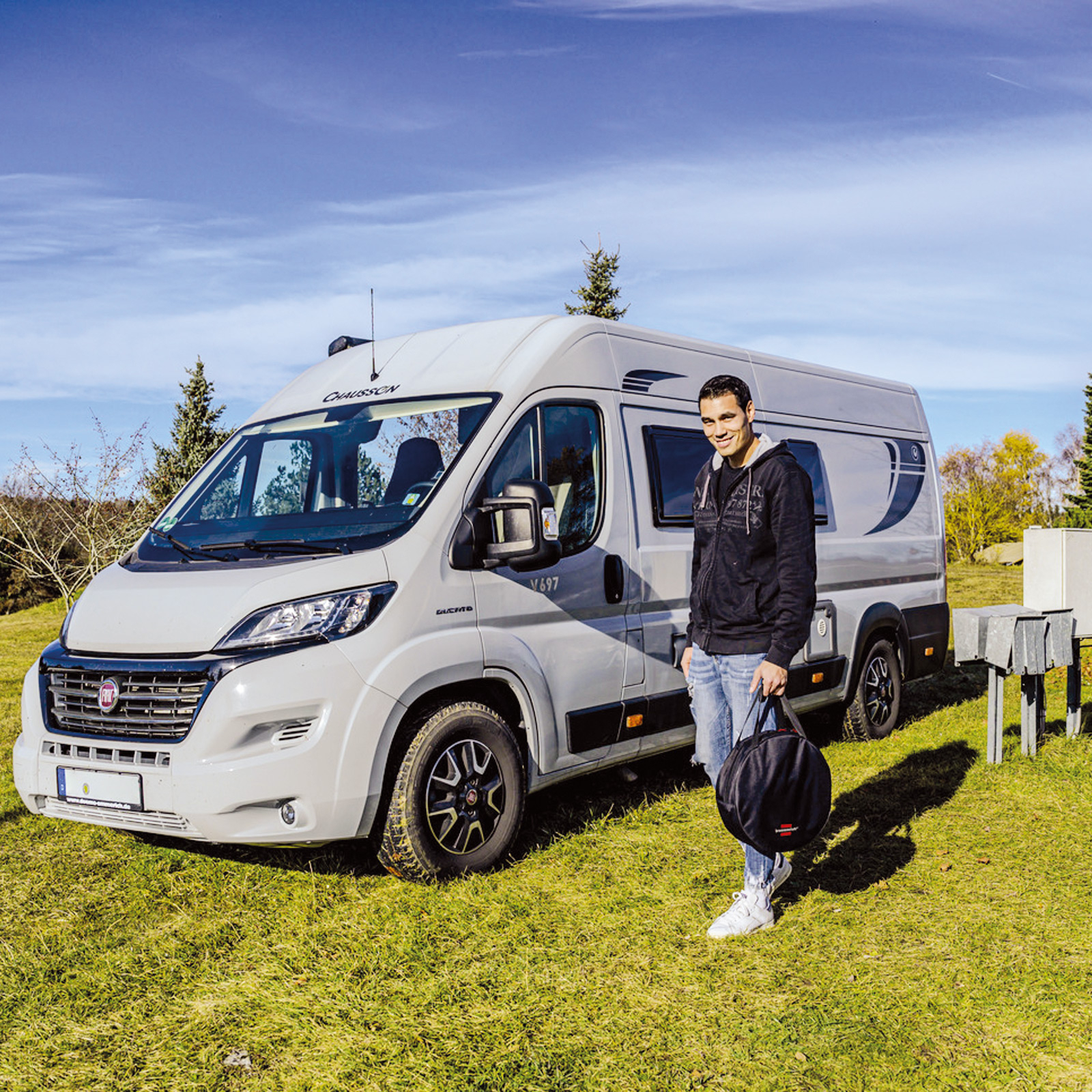 Mężczyzna przed kamperem na trawie, trzyma czarną torbę pod błękitnym niebem.