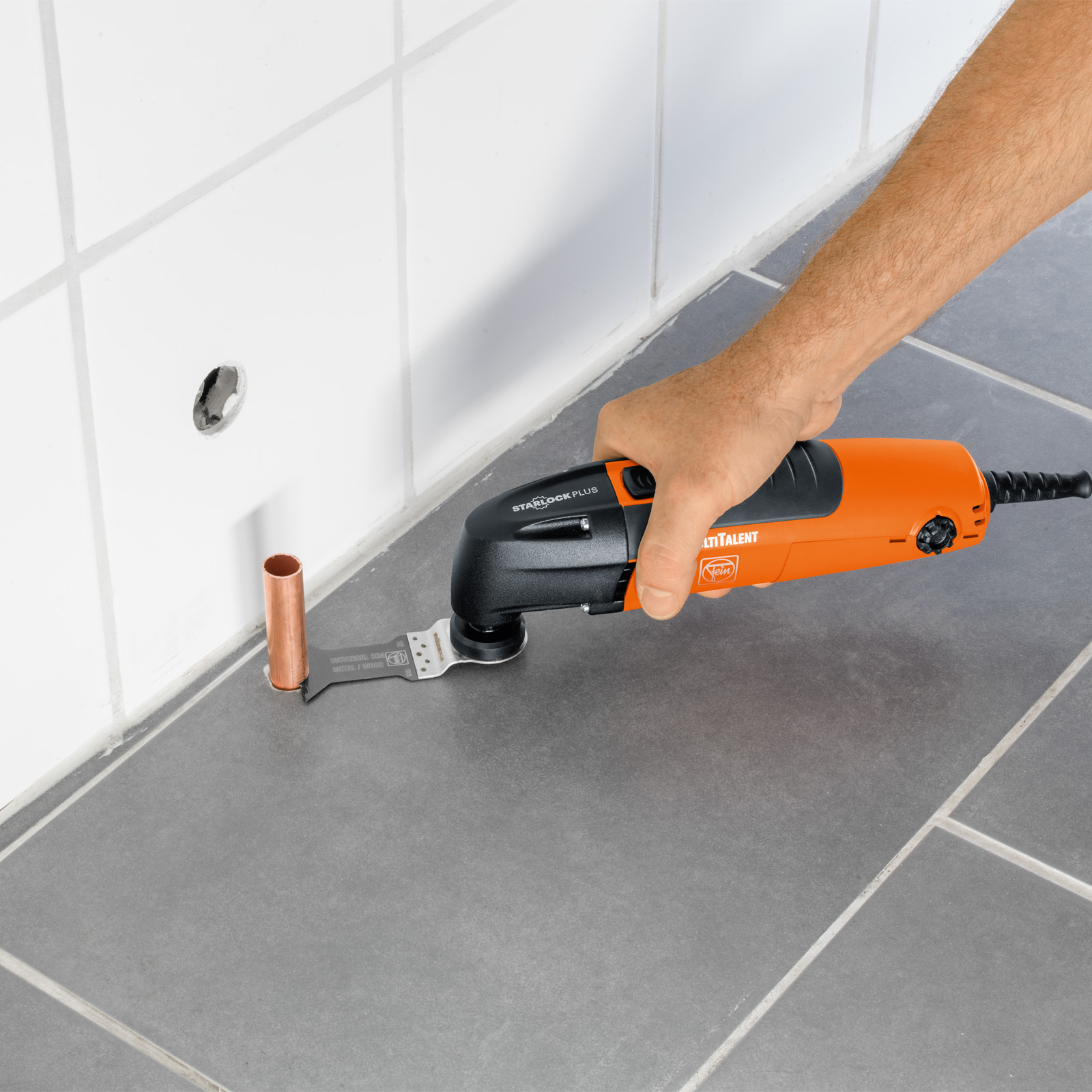 Eine Person benutzt ein orangefarbenes Multitool, um ein Kupferrohr in der Nähe von weißen Fliesen zu schneiden.