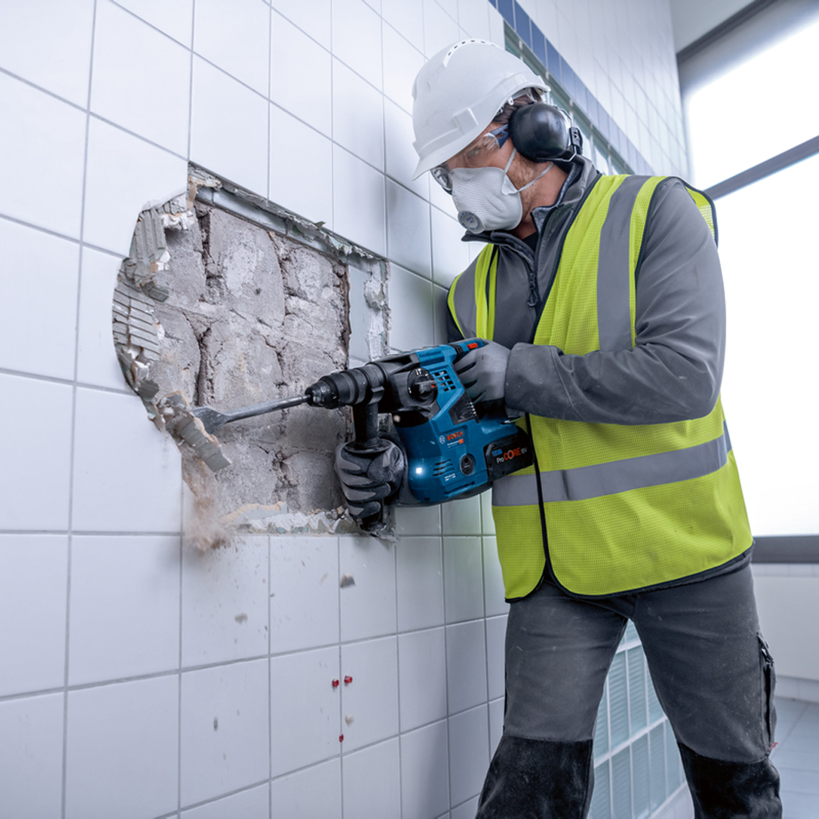 Pracownik budowlany używa niebiesko-czarnej wiertarki udarowej Bosch do rozbiórki ściany.