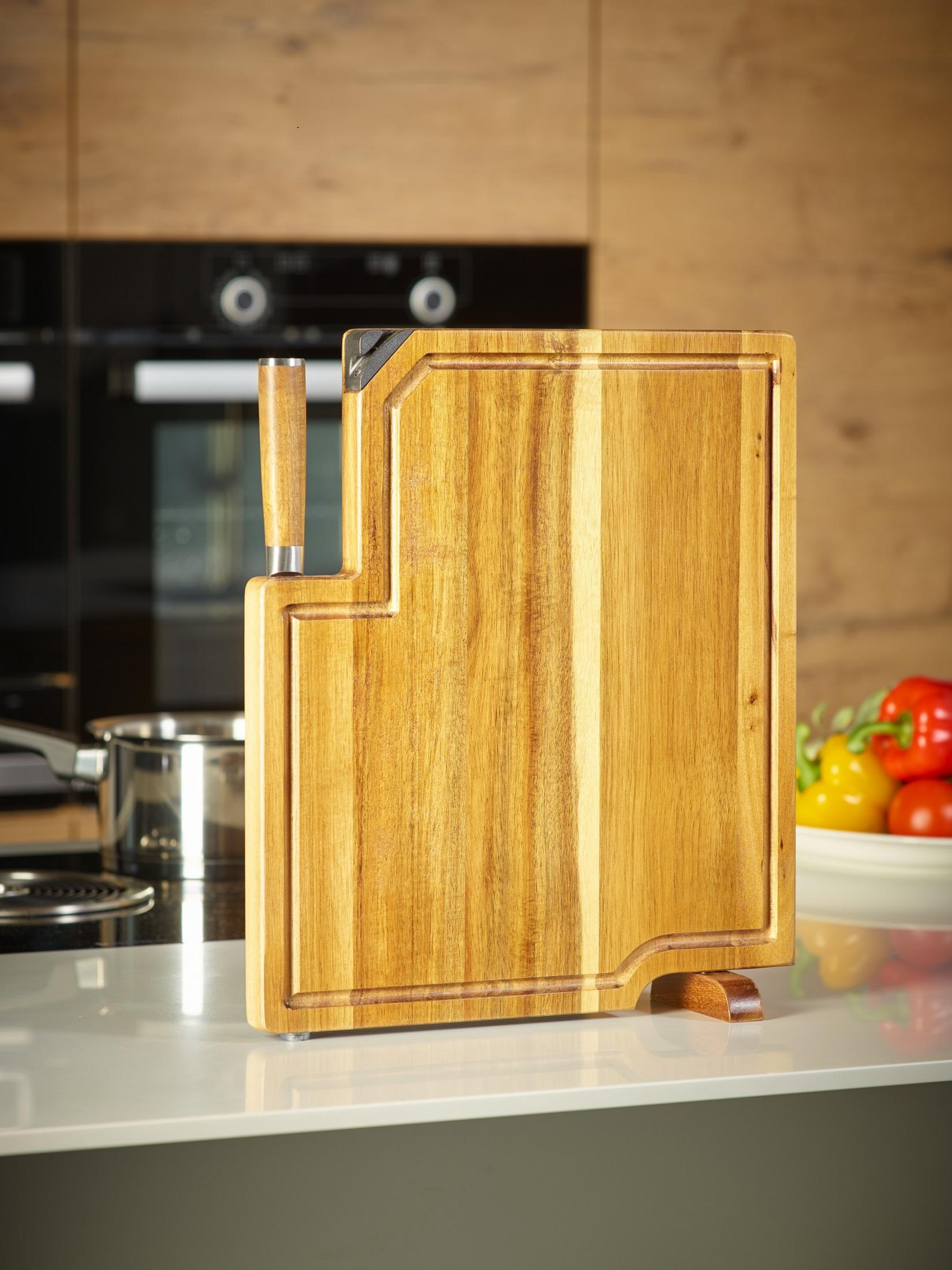 Hölzernes Schneidebrett mit einem Messer, das in einer Küchenumgebung mit Geräten und Gemüse steht.