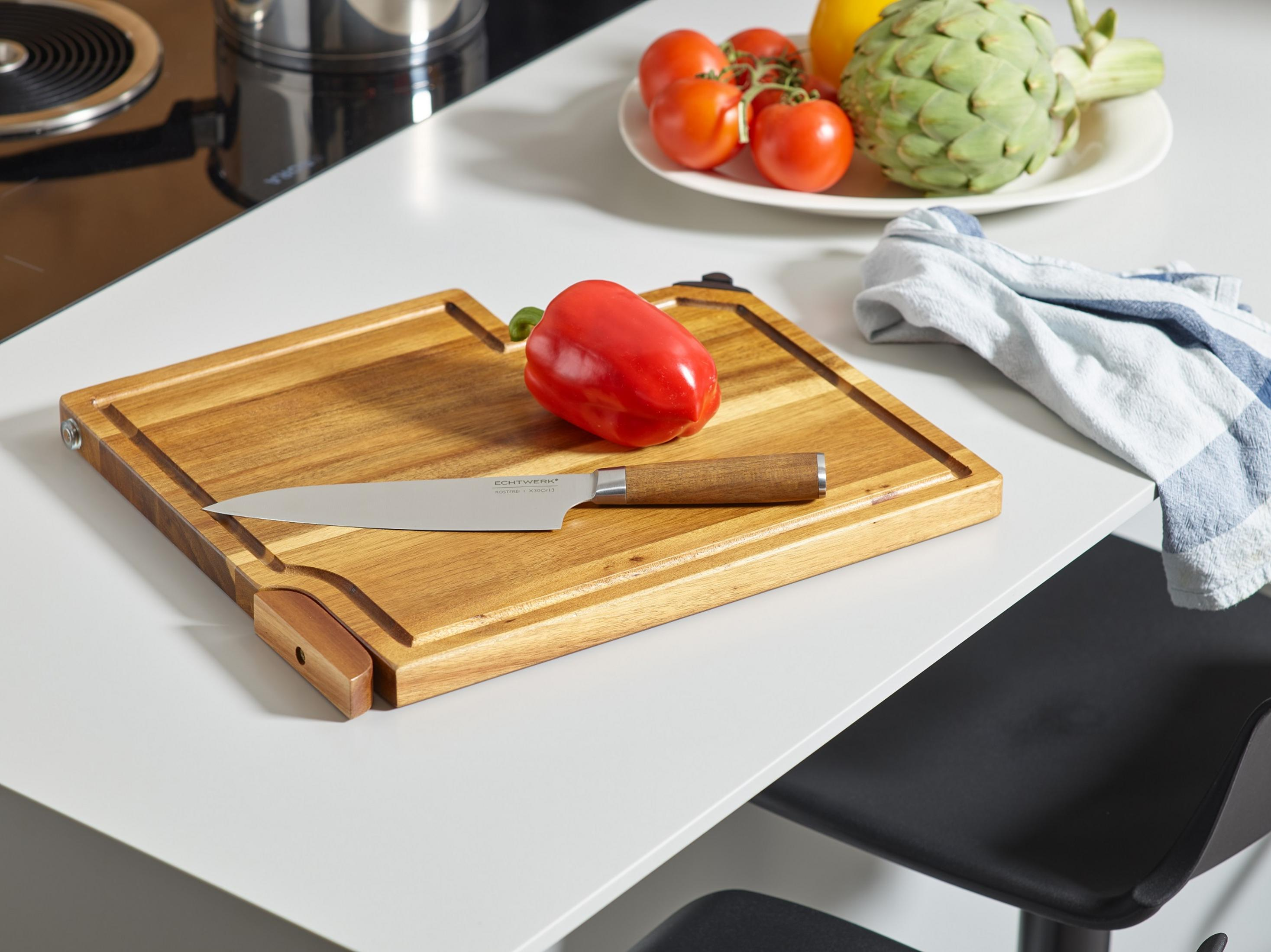 Ein Holzschneidebrett mit Messer und rotem Pfeffer auf einem weißen Tisch mit Tomaten und Artischocke im Hintergrund.