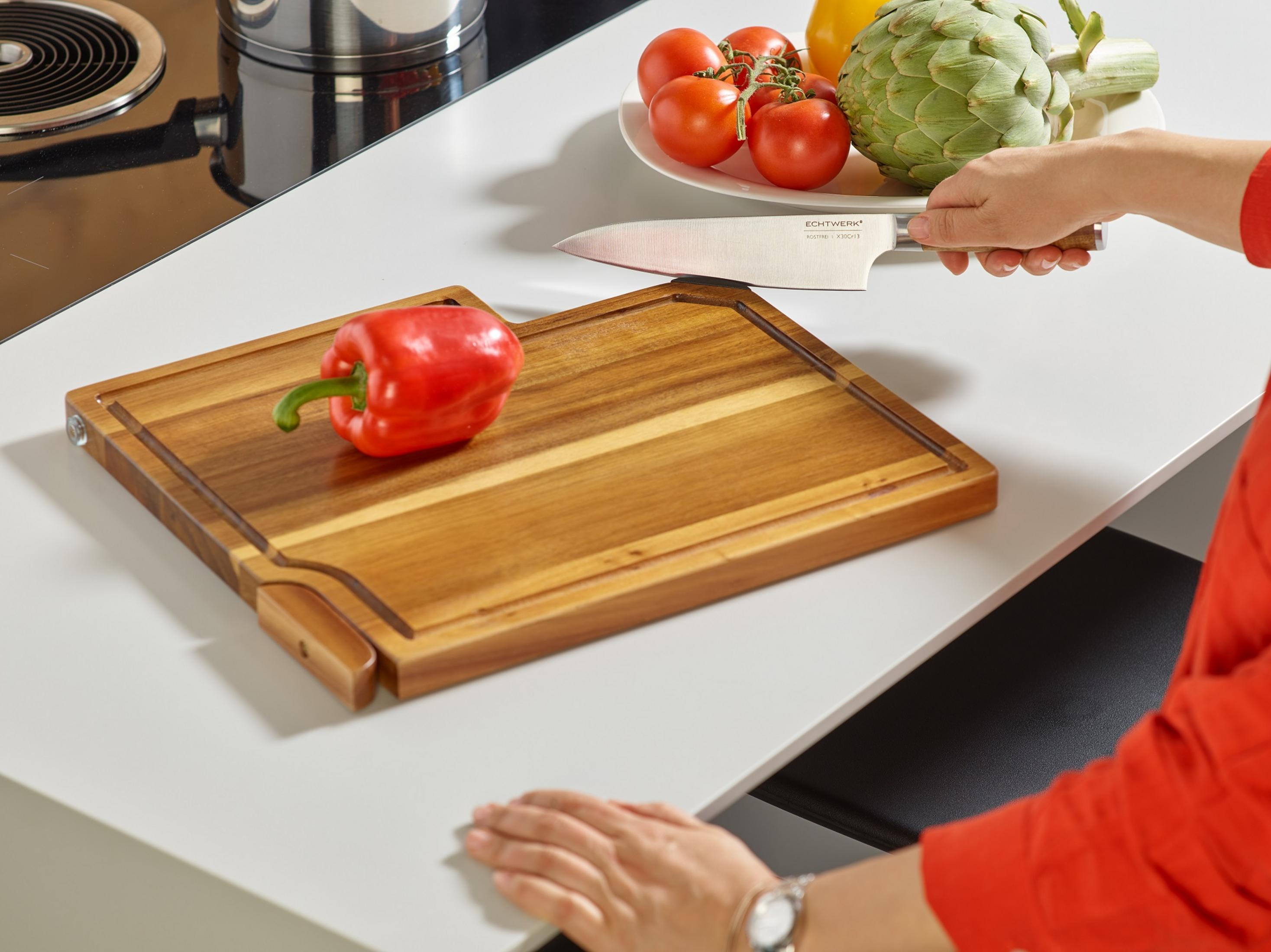 Ein Holzschneidebrett mit rotem Pfeffer und Messer auf einem weißen Tisch, die Hände einer Person und etwas Gemüse im Hintergrund.