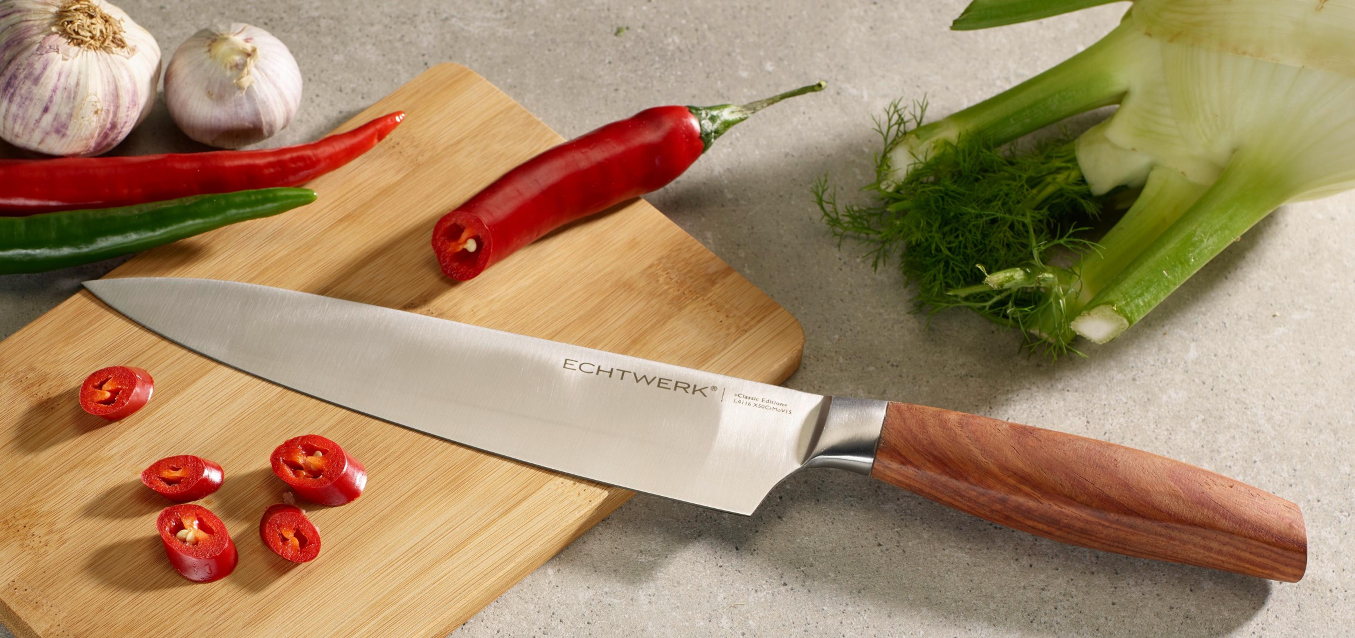 Ein Kochmesser liegt auf einem Schneidebrett mit Paprika, Knoblauch und Fenchel auf einer grauen Oberfläche.