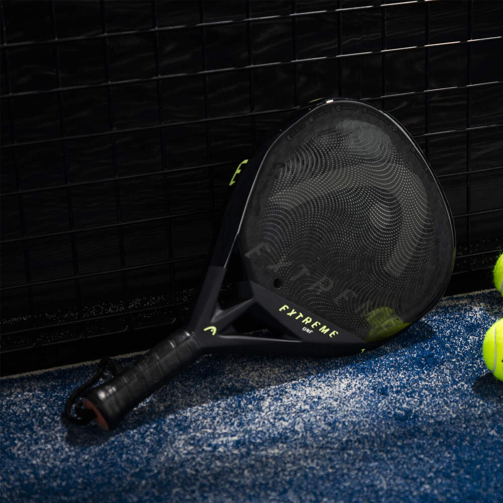 Una racchetta da padel nera con accenti verdi poggia su una superficie blu con palline da tennis nelle vicinanze, su uno sfondo scuro.