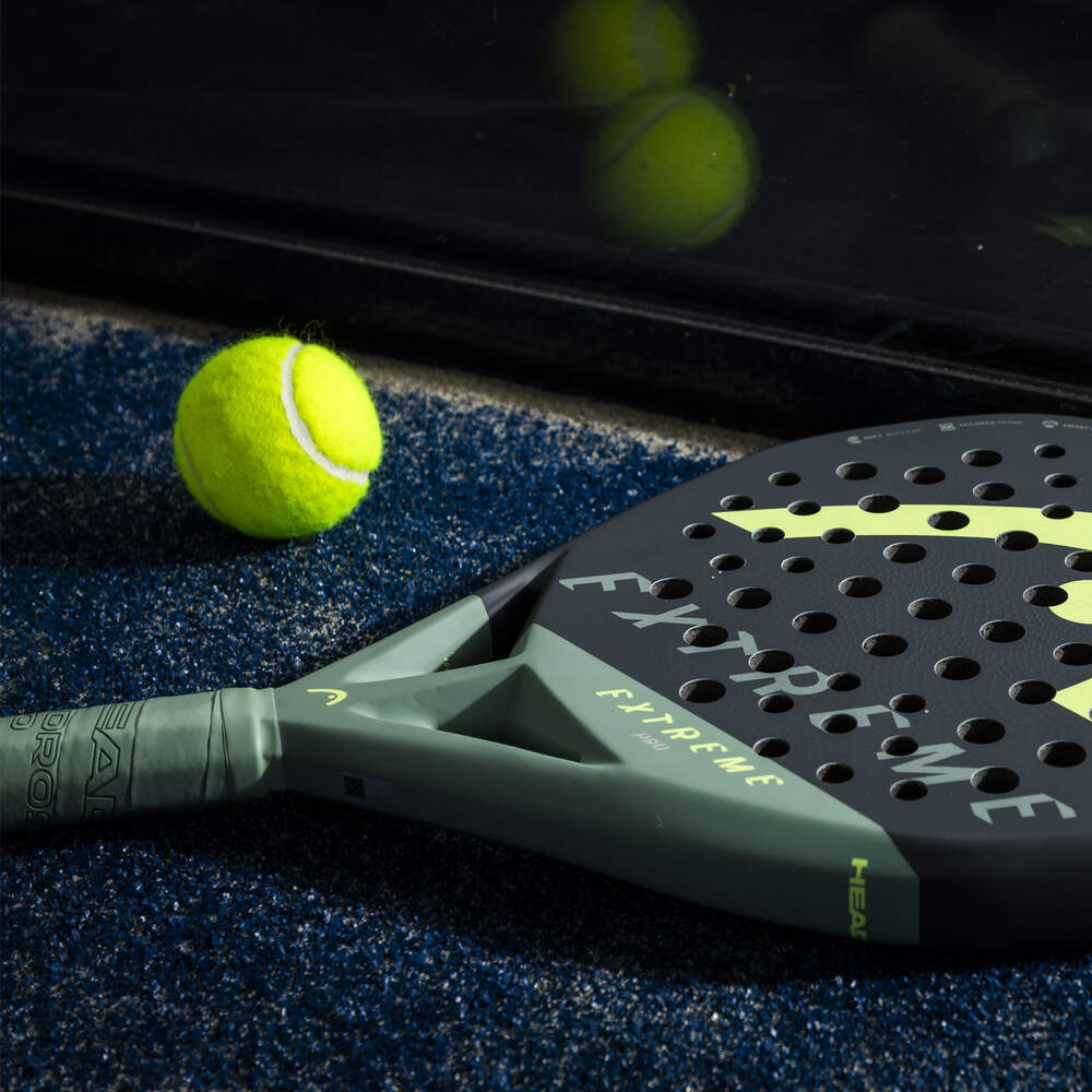 Una racchetta da padel nera e verde con palline da tennis su una superficie blu contro uno sfondo di piastrelle nere.