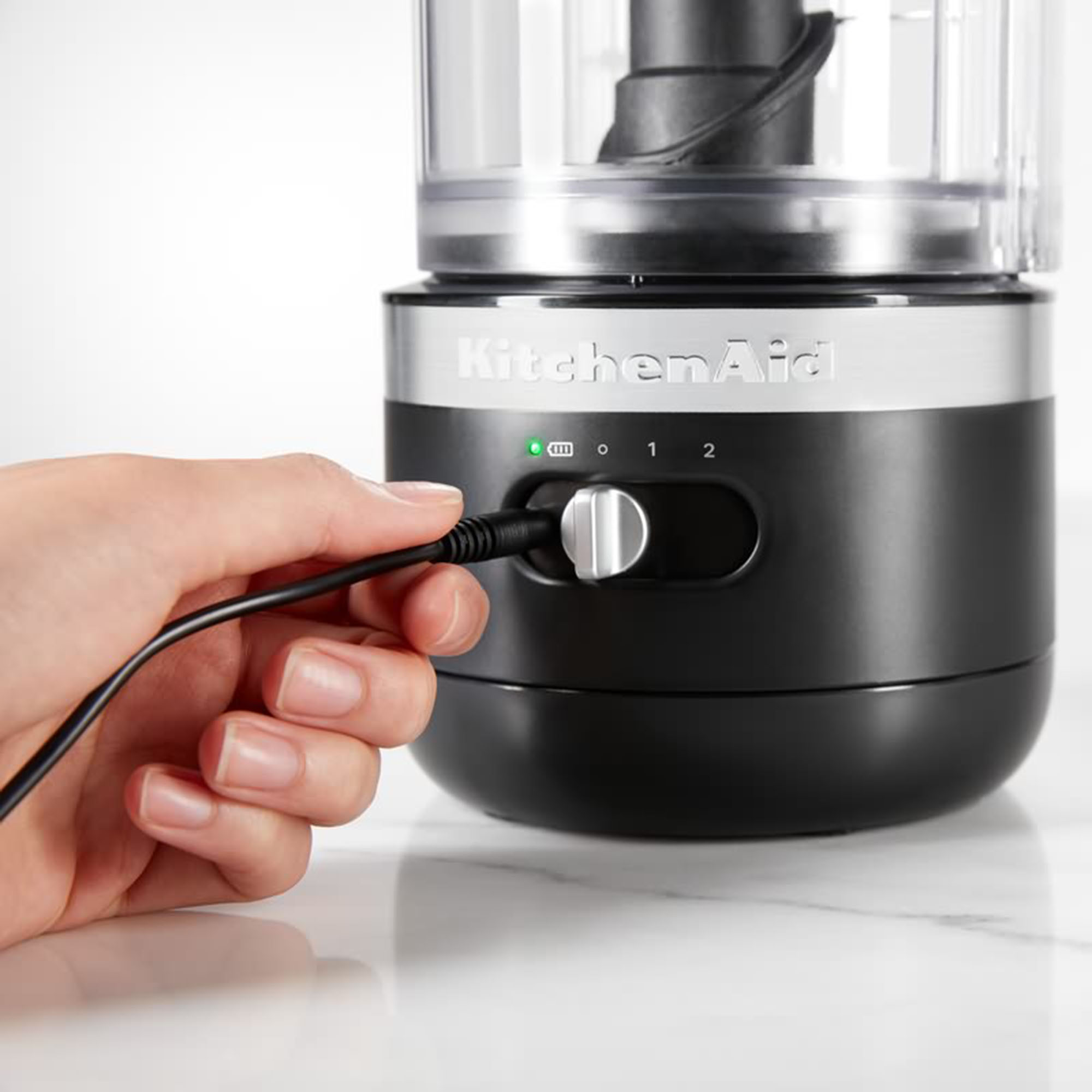 Eine Hand steckt ein schwarzes Stromkabel in eine schwarz-silberne KitchenAid Küchenmaschine auf einer weißen Marmoroberfläche.