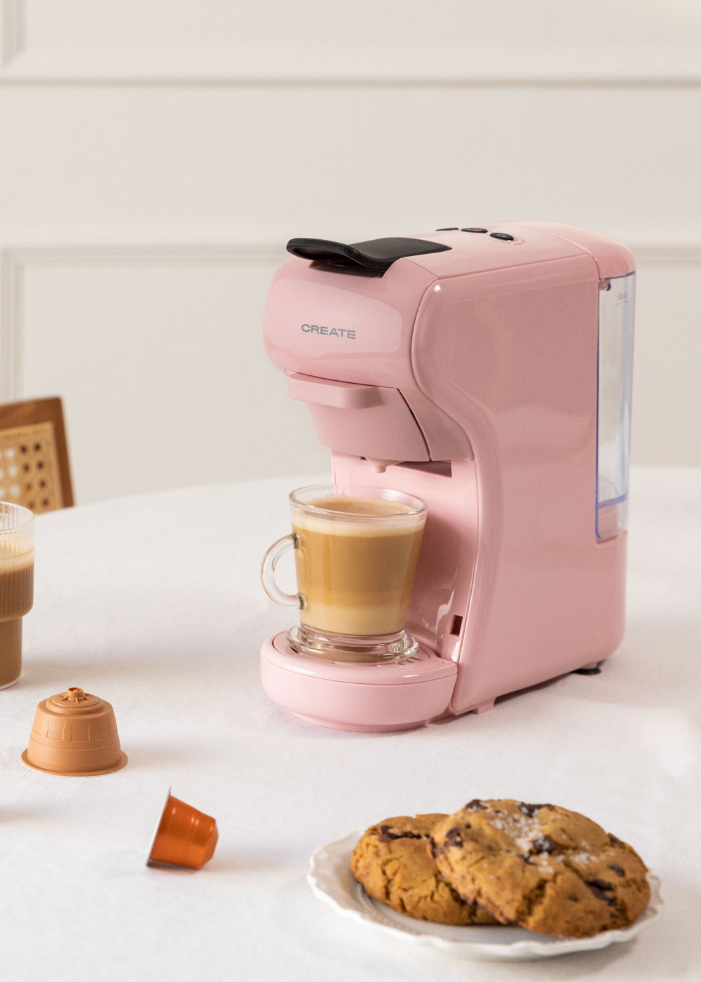 Una cafetera rosa CREATE prepara café en una taza de vidrio, junto a galletas.