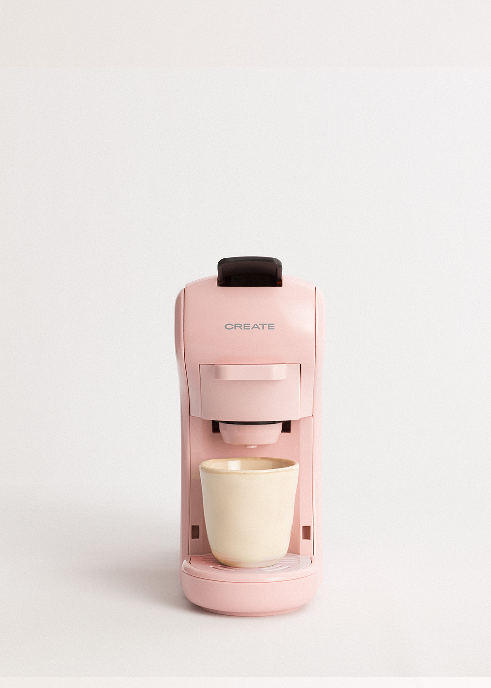 Una cafetera rosa CREATE dispensa café en una taza corta de color crema.