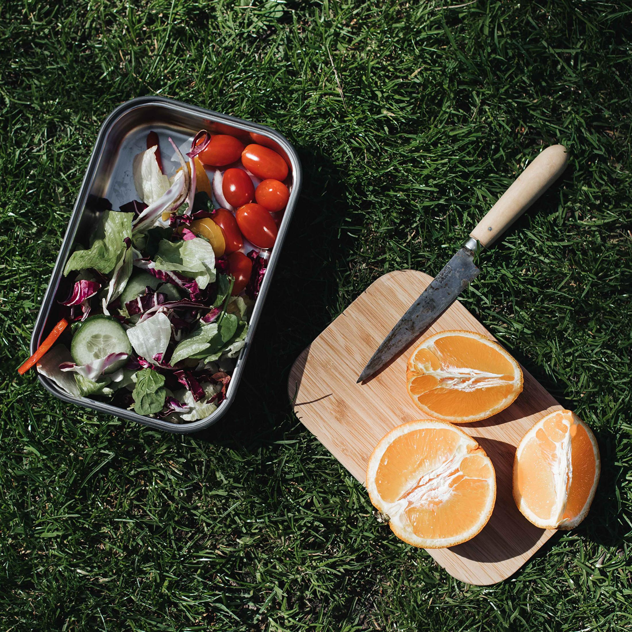 Salat, Tomaten, Orangen und ein Messer liegen auf einem Schneidebrett auf grünem Gras.