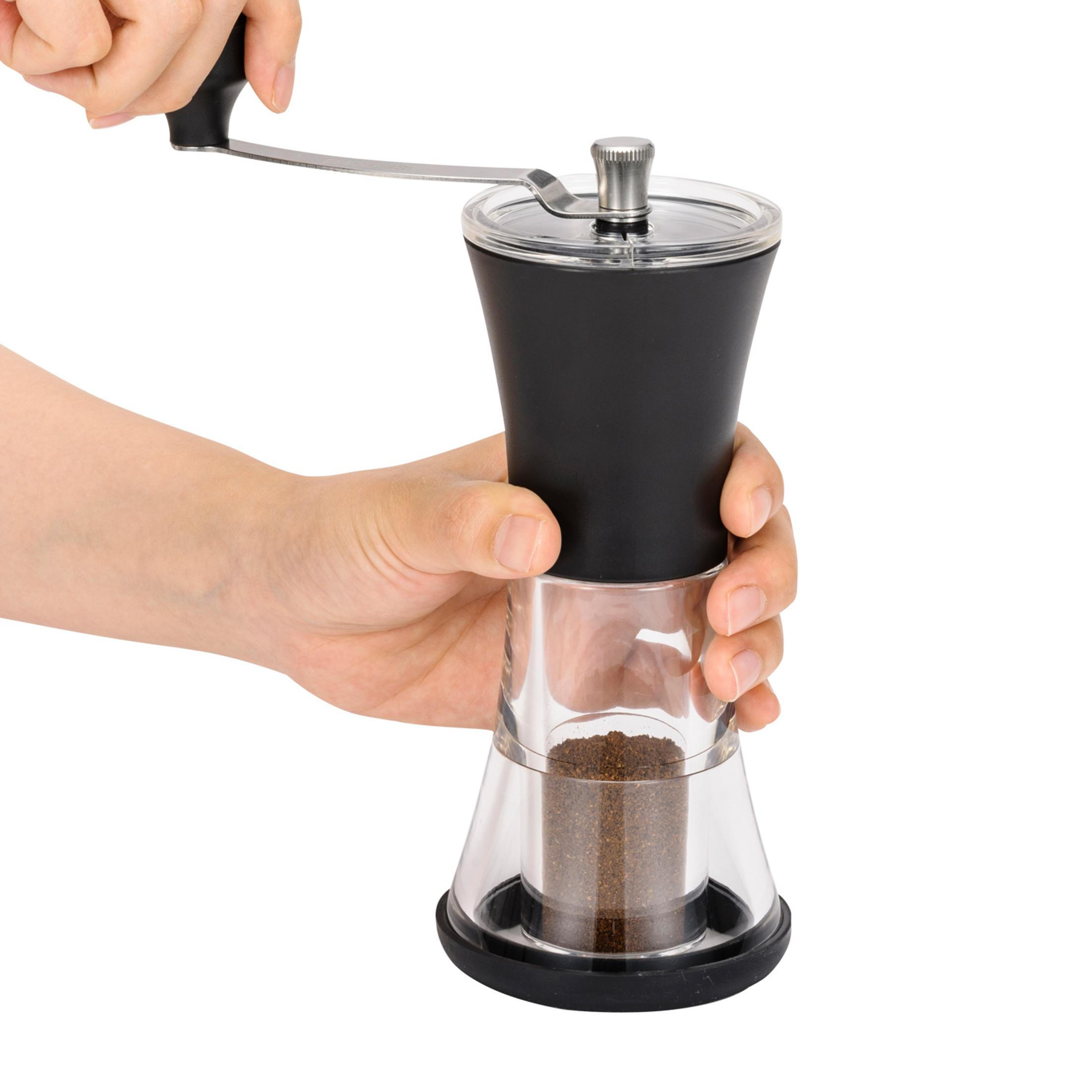 Eine Hand kurbelt eine manuelle Kaffeemühle mit klarem und schwarzem Gehäuse, gefüllt mit gemahlenem Kaffee.