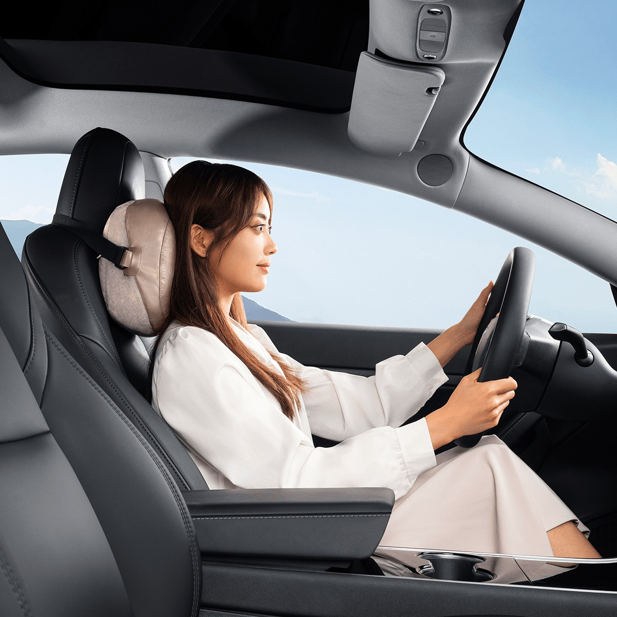 Kobieta prowadzi samochód z poduszką zagłówka. Poduszka jest jasno-brązowa, a wnętrze samochodu jest czarne.
